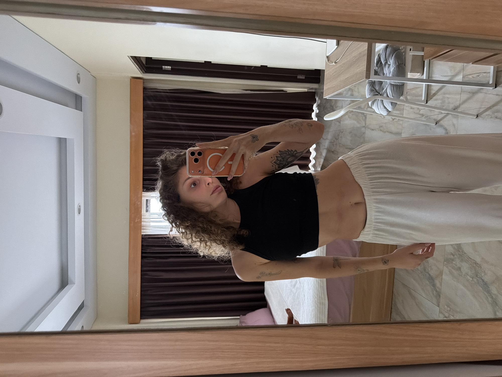 A young woman with curly brown hair takes a mirror selfie in a room in North Kuta, Indonesia. She is wearing a black crop top and white sweatpants. Her left arm, which is visible, has tattoos of a dragonfly and a flower. In the background, a bed with a purple headboard and white bedding is visible, along with dark brown curtains and a modern ceiling with recessed lighting. A white chair with a grey cushion sits next to a wooden desk with a laptop. The floor is tiled with a marble pattern. The woman is holding a mobile phone with an orange case, actively taking the photo.