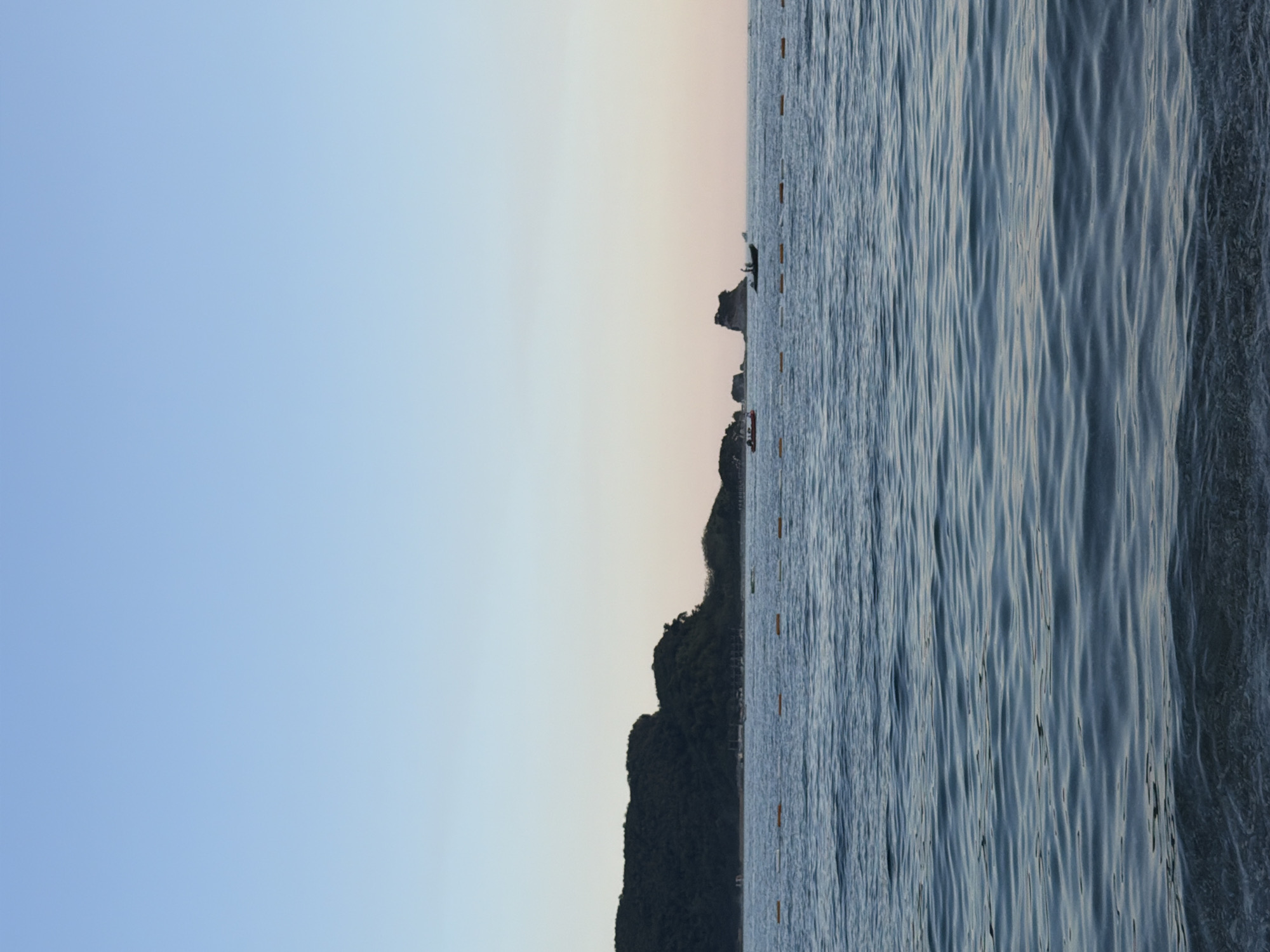 The image shows a serene seascape with a clear sky and calm waters. In the foreground, gentle waves lapped against a pebble beach, with the water appearing a deep blue. Further out, a line of orange buoys marked a boundary in the sea. In the mid-ground, a lush green, tree-covered hill slopes down to the water, suggesting a coastal landscape. The background features a few rocky outcrops rising from the sea, adding a dramatic touch to the horizon. Two small boats are visible on the water, one red and one dark, with a person standing on each. The sky is a soft gradient of light blue, transitioning to a pale pinkish hue near the horizon, indicating either dawn or dusk. This scene is characteristic of the coastline near Budva, Montenegro, known for its beautiful beaches and rocky islands.