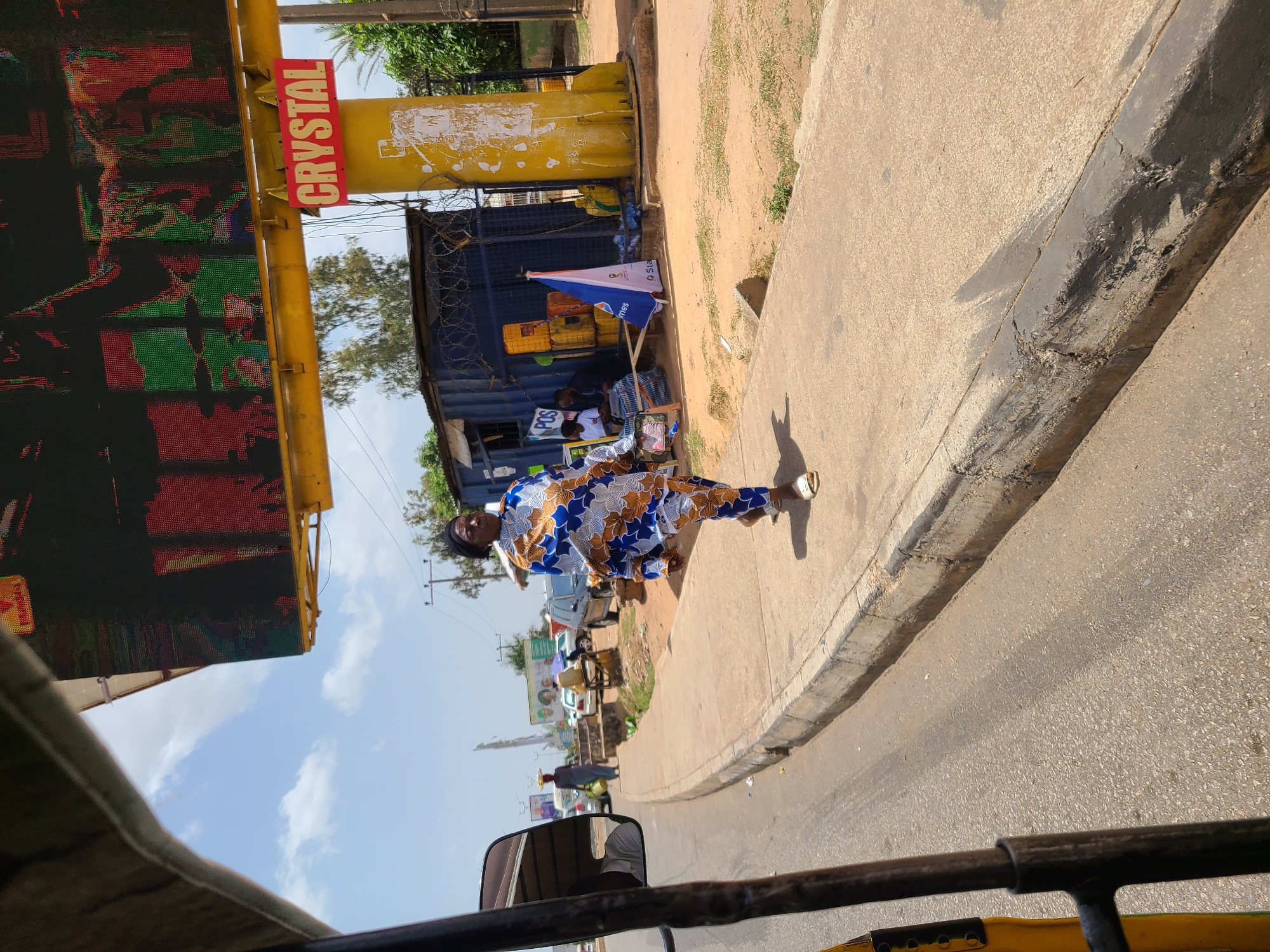 This image captures a vibrant street scene in Jos, Nigeria, observed from inside a moving vehicle, with part of its frame visible in the foreground. The time of day is bright and sunny, indicated by the clear blue sky with scattered clouds and distinct shadows.
In the center, a woman is walking on a paved sidewalk. She is dressed in a distinctive, colorful patterned outfit in shades of blue, white, and brown, and wears light-colored sandals.
To the left, a large LED billboard, framed in yellow, displays dynamic, pixelated red and green graphics. Mounted above it is a red sign with yellow lettering that reads "CRYSTAL". Adjacent to the billboard structure is a large yellow pipe or pole.
Further down the sidewalk, a makeshift stall operates out of a blue container or structure. A white and blue umbrella provides shade, bearing partial text "Sta" and "mes" (likely part of "StarTimes" or similar branding). The letters "POS" are clearly visible on the stall, suggesting a point-of-sale service. Several people are gathered near this stall.
The street itself is an asphalt road separated from the sidewalk by a concrete curb. Beyond the immediate foreground, other vehicles and distant buildings are visible, adding to the urban context. The scene portrays a typical bustling commercial or residential street environment.