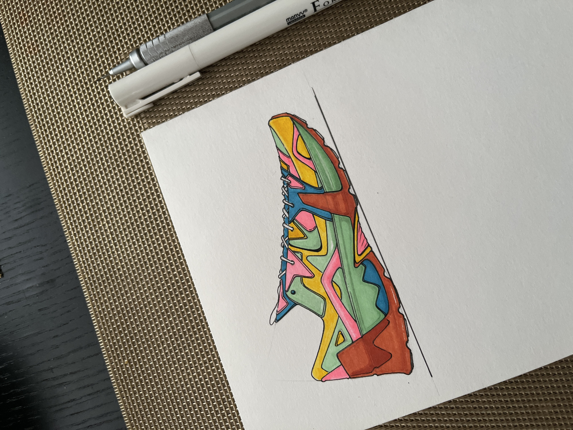 The image shows a colorful drawing of a sneaker shoe on a piece of white paper. Two pens, one white and one gray, are placed at the top of the paper. The paper and pens sit on top of a gold-colored woven surface. To the left of the woven surface is a black surface. The drawing is the main subject, and the pens and surfaces provide context to the image. The location and city are unknown.
