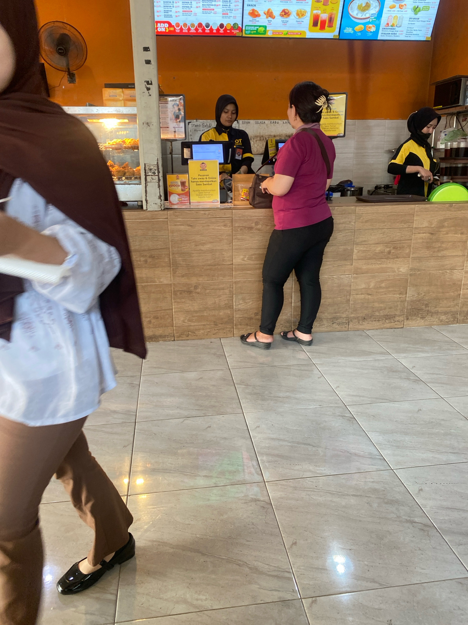 The image captures a scene inside a food establishment, likely a fast-food restaurant. A woman wearing a maroon hijab and a pink shirt, with black leggings and sandals, stands at a counter, seemingly waiting to order or pay. To her right, another woman in a yellow and black uniform, also wearing a hijab, works behind the counter, possibly as an employee. In the background, a display case filled with fried chicken is visible, along with menu boards and various food items. The restaurant appears to be situated in Semarang, Indonesia, indicated by the Indonesian text on the menu and signs. The floor is tiled with a marble-like pattern. The overall atmosphere suggests a busy, casual dining environment.