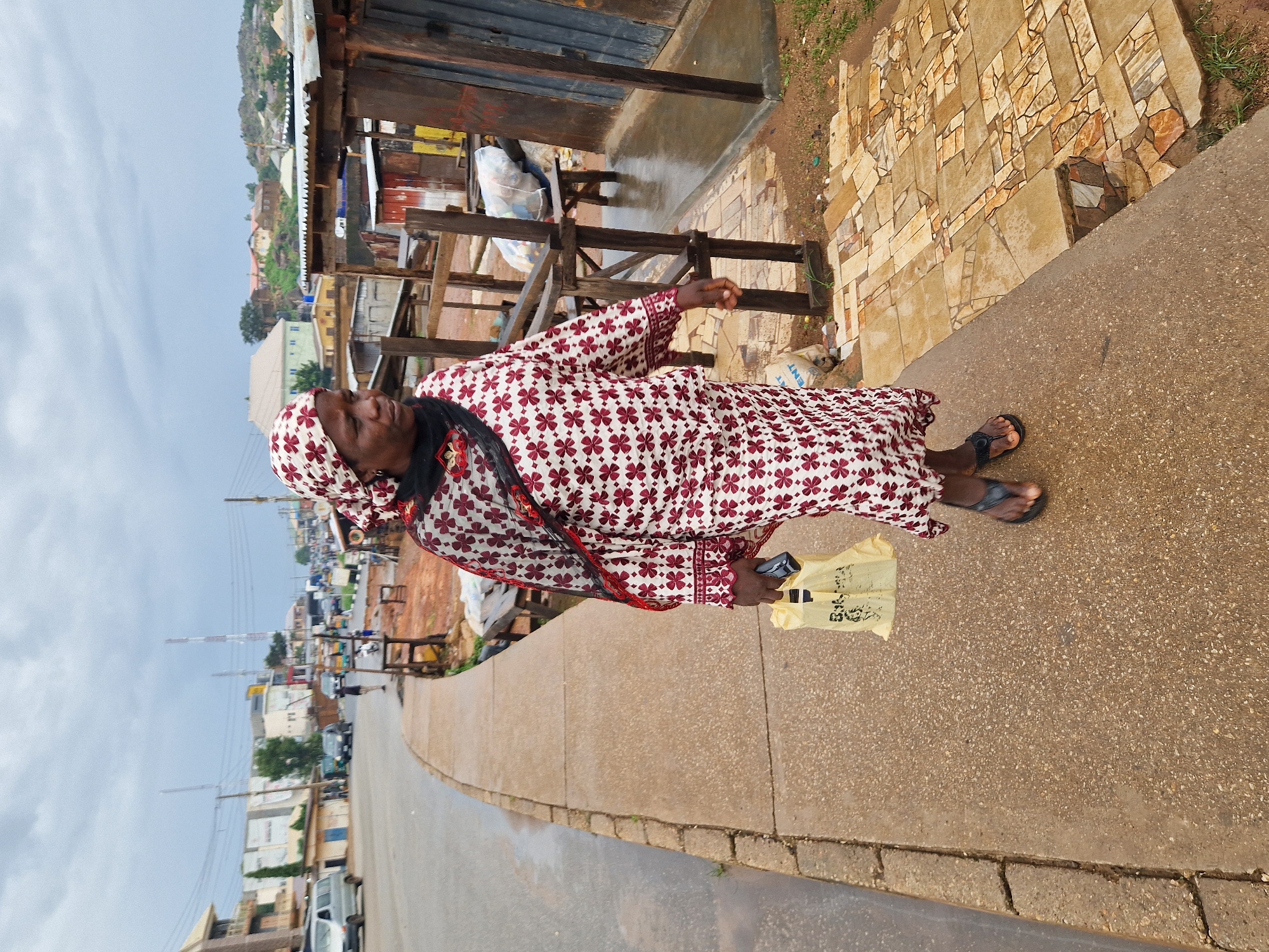 A full-body, outdoor, eye-level shot shows an adult woman walking on a sidewalk in Kabong, Nigeria. The woman is wearing a maroon and white patterned dress with a matching headscarf and a black scarf around her neck. She is holding a yellow plastic bag in her left hand and a dark-colored object, possibly a cell phone, in her right hand. She is wearing black flip-flops. The sidewalk is made of concrete with a rough texture. To the right of the sidewalk, there are several wooden market stalls with corrugated metal roofs. The ground around the stalls is dirt and gravel, with some areas paved with irregular-shaped stones. In the background, there is a street with vehicles, buildings, and utility poles, suggesting an urban environment. The sky is overcast, and the ground appears wet, indicating recent rain. The overall scene depicts a typical street scene in a Nigerian city.