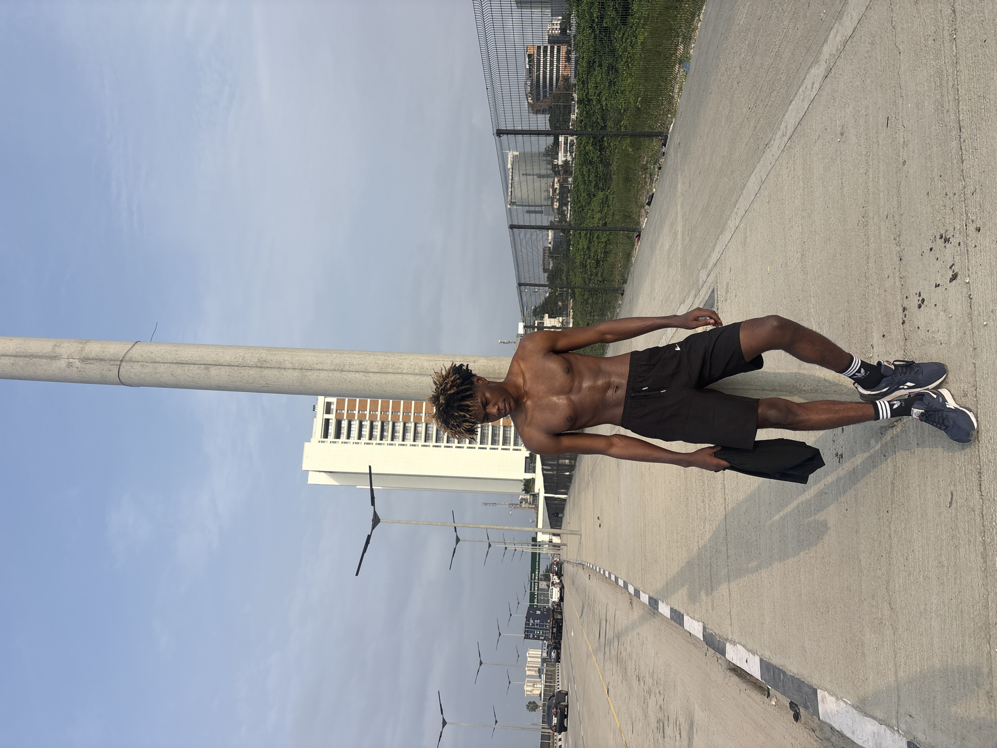 In Alaguntan, Nigeria, a young Black male stands shirtless, leaning against a concrete lamppost on a pedestrian walkway. He is wearing black athletic shorts, black and white striped socks, and dark blue athletic shoes. He holds a dark t-shirt in his left hand. The walkway runs alongside a multi-lane highway with traffic visible in the distance. Several modern streetlights line the highway. In the background, a tall, modern white building with multiple floors and balconies stands prominently. Beyond the highway, a chain-link fence encloses a grassy area, with more buildings and urban landscape visible further in the distance. The sky is a light blue with scattered clouds, and the sun casts long shadows on the walkway.