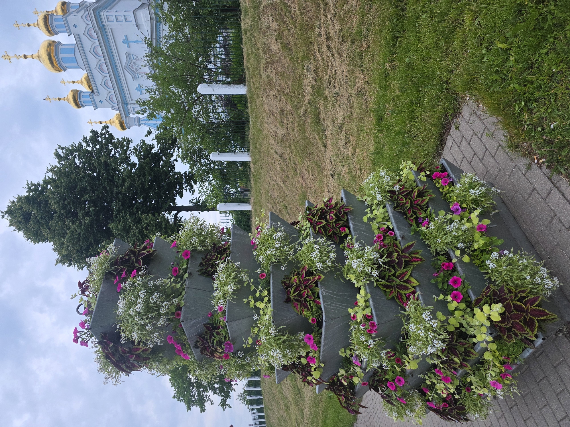This image depicts an outdoor scene featuring a large, tiered planter filled with various flowers and foliage in the foreground, with a church visible in the background. The location is in the city of Unknown, Unknown.
In the immediate foreground, a multi-level planter, constructed from gray, angular sections, is situated on a gray paver brick walkway. The planter is filled with a mix of plants, including bright pink petunia-like flowers, small white trailing flowers (possibly alyssum), and several varieties of coleus or similar foliage plants with variegated green, red, and deep purple leaves. Some lighter green trailing plants are also present. The planter rises vertically, appearing as a stack of seven or more offset square or trapezoidal tiers.
To the right of the planter and walkway, a grassy area slopes upwards. The grass on the slope appears dry and matted, while the grass on the flatter ground near the walkway is greener. Along the crest of the slope, a dark metal fence with vertical pickets is visible, punctuated by several white, rectangular stone or concrete posts. Behind the fence, a line of green trees and shrubs forms a natural barrier. A large, dense deciduous tree stands prominently on the left side of the frame, partially obscuring the background.
In the far background, positioned on top of the hill and behind the trees, is a light-colored building, likely a church or cathedral, characterized by its multiple golden onion domes, each topped with a golden Orthodox cross. The building features white and light blue decorative elements, including arches and ornate detailing along its roofline. At least five golden domes are visible.
The sky overhead is overcast, displaying varying shades of gray and white, suggesting a cloudy day. There are no people visible in the image.