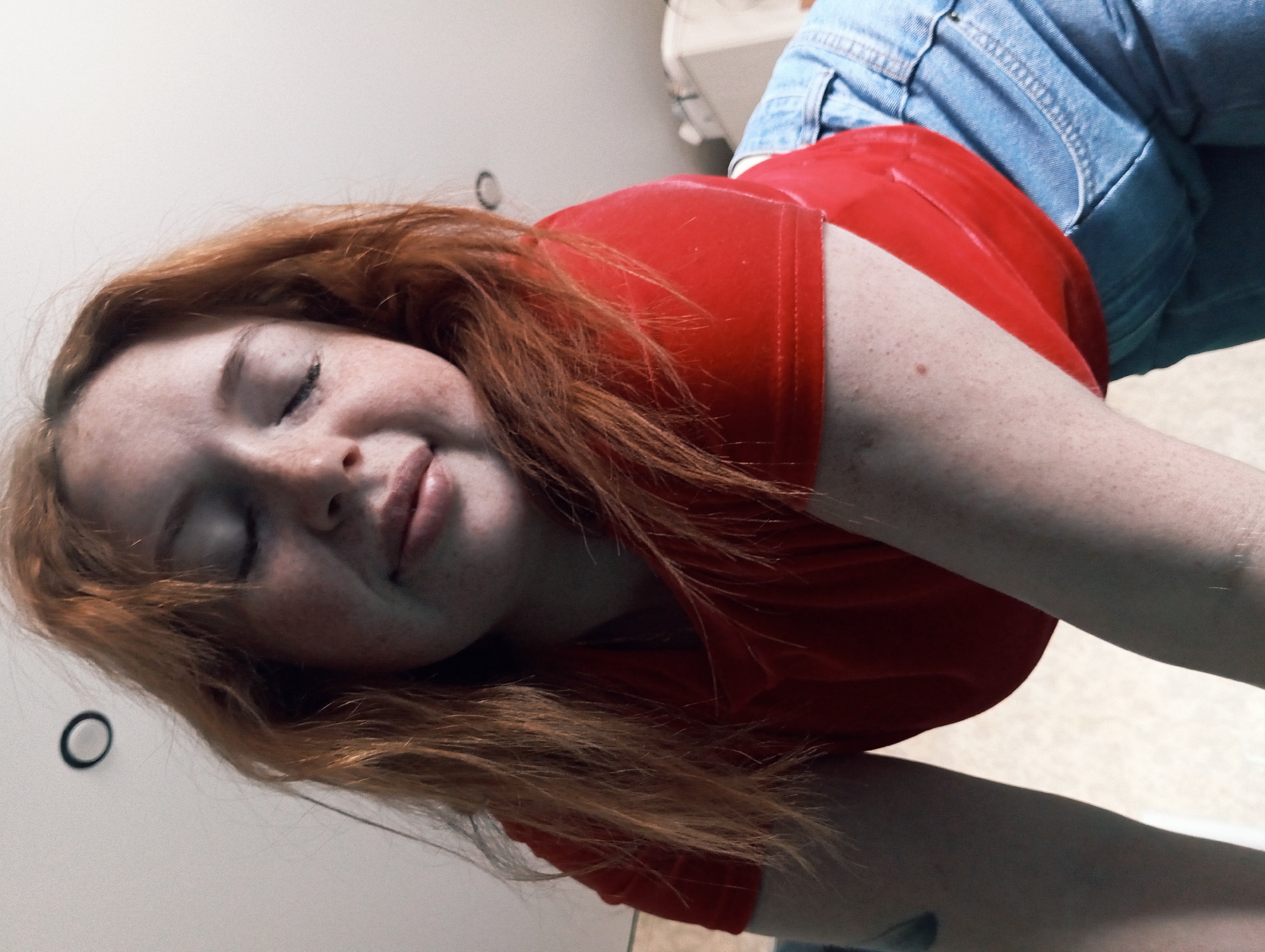 A fair-skinned, freckled person with red hair is seen lying down. The person is wearing a red shirt and blue jeans. The background appears to be a plain white wall and a light-colored floor. There is a small circular object on the wall. The location is not identifiable as Yurgamysh, Russia from the visual data provided.