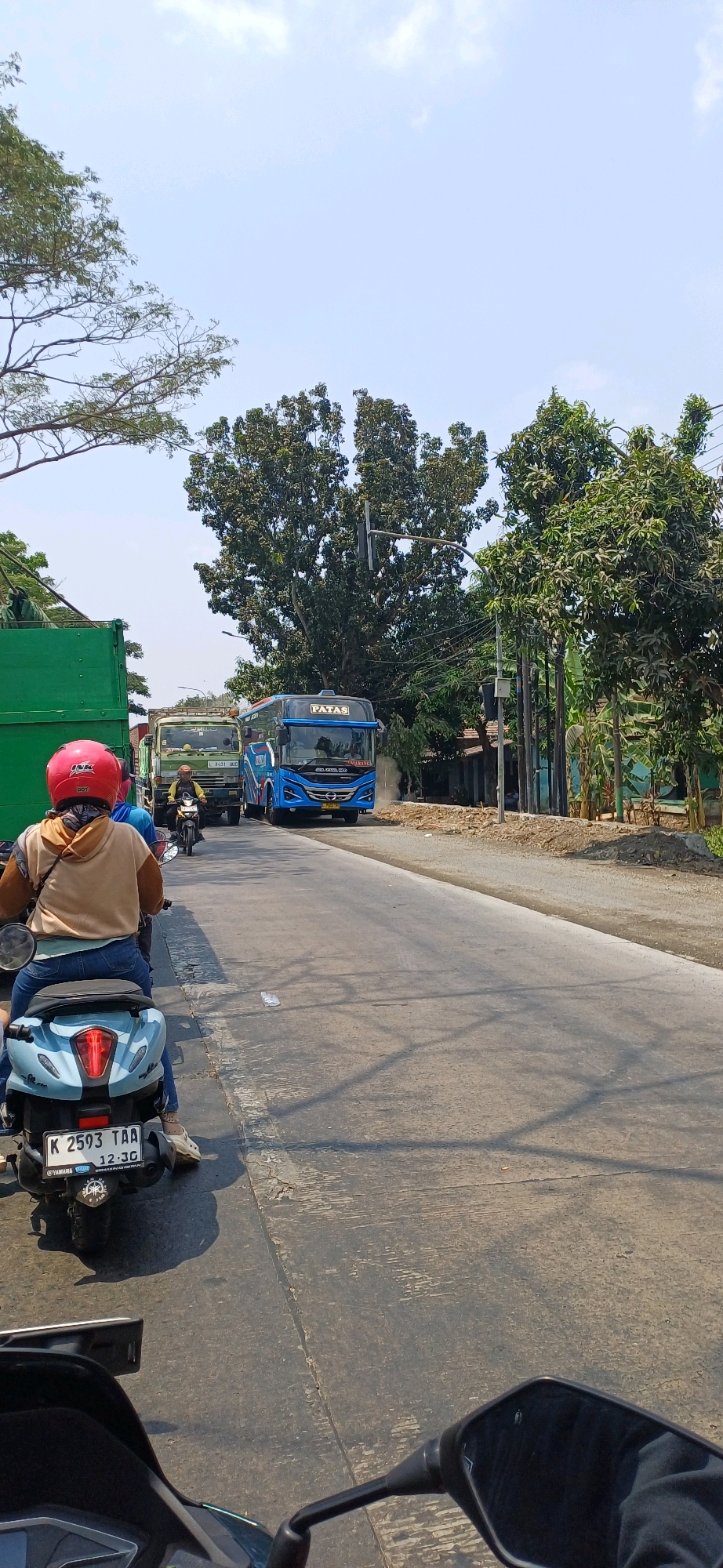 The image captures a street scene in Kecamatan Pati, Indonesia, on a sunny day with a clear sky. In the foreground, a person on a light blue scooter wears a tan hoodie and blue jeans, with a red helmet. The scooter has a license plate reading "K 2593 TAA 12-30". The road ahead is paved and shows signs of wear.
Traffic is present, with a blue bus prominently visible in the middle ground, displaying the name "PATAS" on its front. The bus is flanked by other vehicles, including a green truck to its left, which has a license plate that is partially obscured but appears to read "L 8431 JHC". Behind the green truck, another vehicle is visible. Further back, a person on a motorcycle is seen riding in the same direction as the scooter in the foreground.
The environment is characterized by lush green trees lining the right side of the road and scattered vegetation on the left. Buildings are visible in the background on the right, with a power pole and electrical wires crossing the scene. The road appears to be a main thoroughfare, suggesting it's a commercial or residential area within the town. The overall atmosphere is that of a typical day with moderate traffic flow.