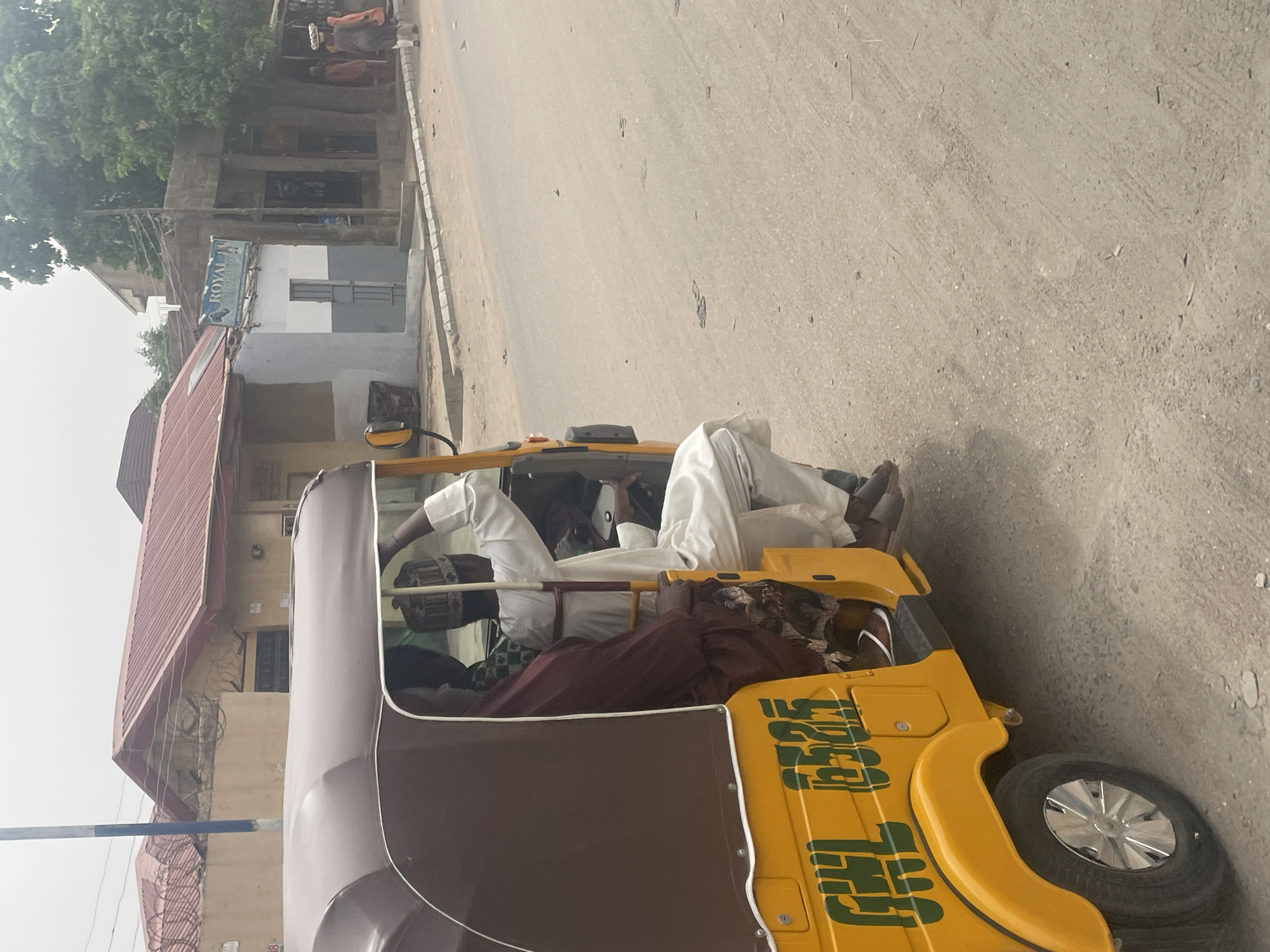 A yellow and brown auto-rickshaw sits on a street in Unknown, Unknown. Inside the vehicle, two men are dressed in white robes and hats. The street appears paved with sand. Buildings are visible behind the auto-rickshaw.