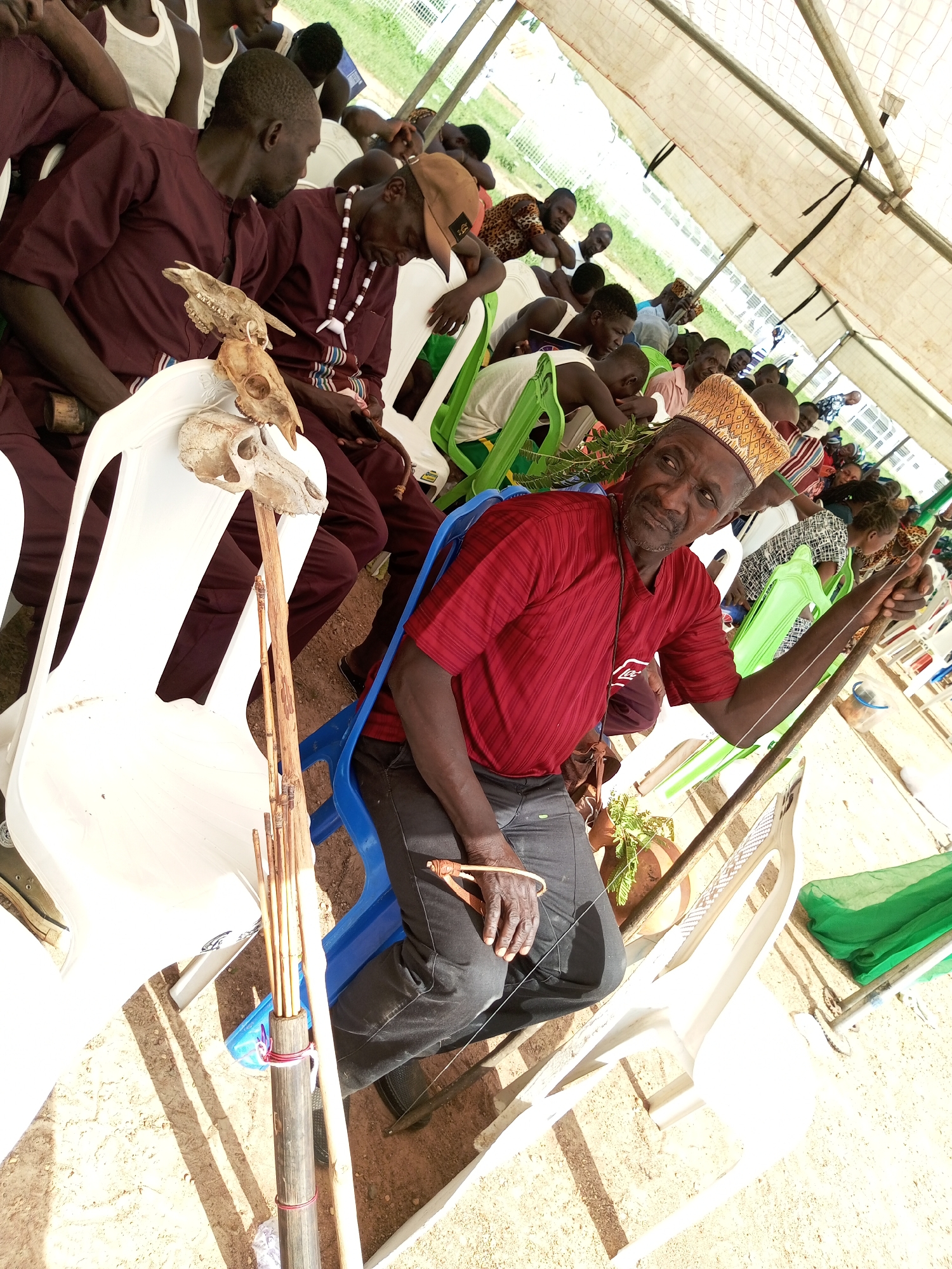 In Takau, Nigeria, a gathering unfolds under a white tent, as evidenced by the tent ceiling overhead. Several individuals sit on plastic chairs, a mix of white, blue, and green. In the foreground, a man wearing a red shirt and a patterned hat sits on a blue chair, holding a long, stringed instrument, possibly musical. Next to him is a white chair with a staff leaning against it, adorned with animal skulls at the top. Other people in the background are dressed in various attires, some in maroon shirts and others in casual wear. The ground appears to be sandy, with shadows cast by the tent structure and people.