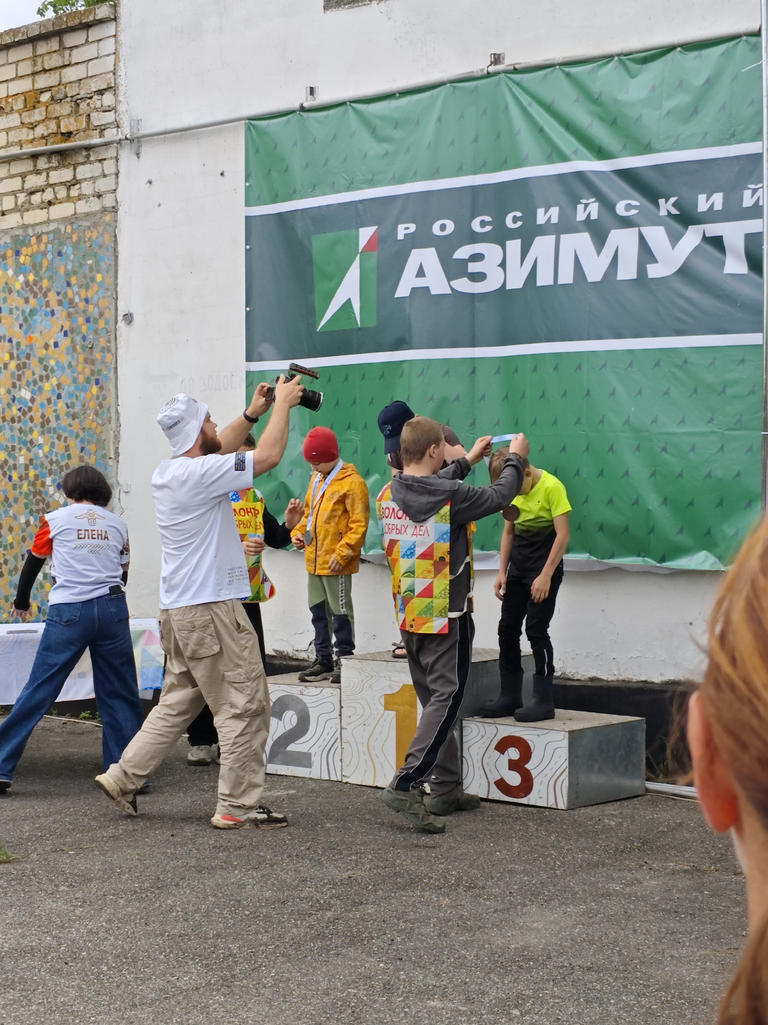 A photo taken in Новоекатериновская, Russia, shows a group of people near a building with a banner. A white banner with the Russian words "РОССИЙСКИЙ АЗИМУТ" is on the wall. In front of it, a podium with the numbers "1", "2", and "3" is visible. Several people are standing on or around the podium. One person in a white hat and khaki pants is taking a photo with a camera. A child in an orange jacket and a red hat stands on the podium. Another individual, wearing a vest, appears to be placing a medal around the neck of a child in a neon green shirt standing on the "3" block. A woman wearing a "ЕЛЕНА" t-shirt is standing near the podium. The setting appears to be an outdoor event or competition.