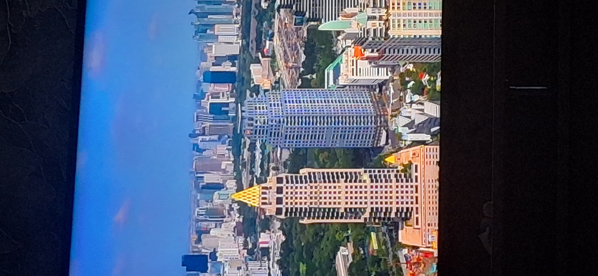 The image is an aerial view of a densely populated city skyline under a bright blue sky with scattered clouds. The foreground features several prominent skyscrapers, with the most striking being a tall, multi-tiered building with a golden, pyramid-shaped apex. To its right is another tall skyscraper with a facade of blue and white elements. In the background, a vast expanse of buildings of varying heights and architectural styles stretches to the horizon, interspersed with patches of green trees. The lower portion of the image shows a dark, out-of-focus area, possibly indicating the presence of furniture or the edge of a screen. The scene depicts a bustling urban environment, likely a major metropolitan area, characterized by its modern architecture and significant infrastructure. The location is not Bila Tserkva, Ukraine.