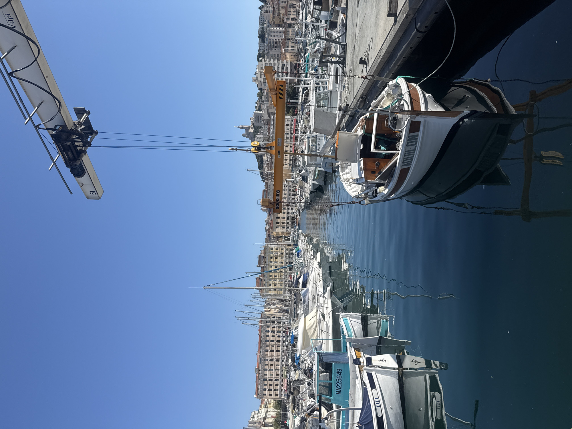 The image depicts a harbor scene under a clear blue sky. In the foreground, a white boat with brown trim named "AQUILON" is suspended by a yellow crane, its reflection visible in the calm water. Several other boats are docked along a concrete pier to the right. In the middle ground, a row of white sailboats with tall masts are moored, their reflections also visible in the water. Further back, a city skyline with buildings of varying heights and colors forms the backdrop, with a large church or basilica visible on a hill overlooking the city. A large industrial crane with a "5t" marking is positioned in the upper left quadrant of the image, with cables hanging down. The overall scene suggests a working harbor in a coastal city.