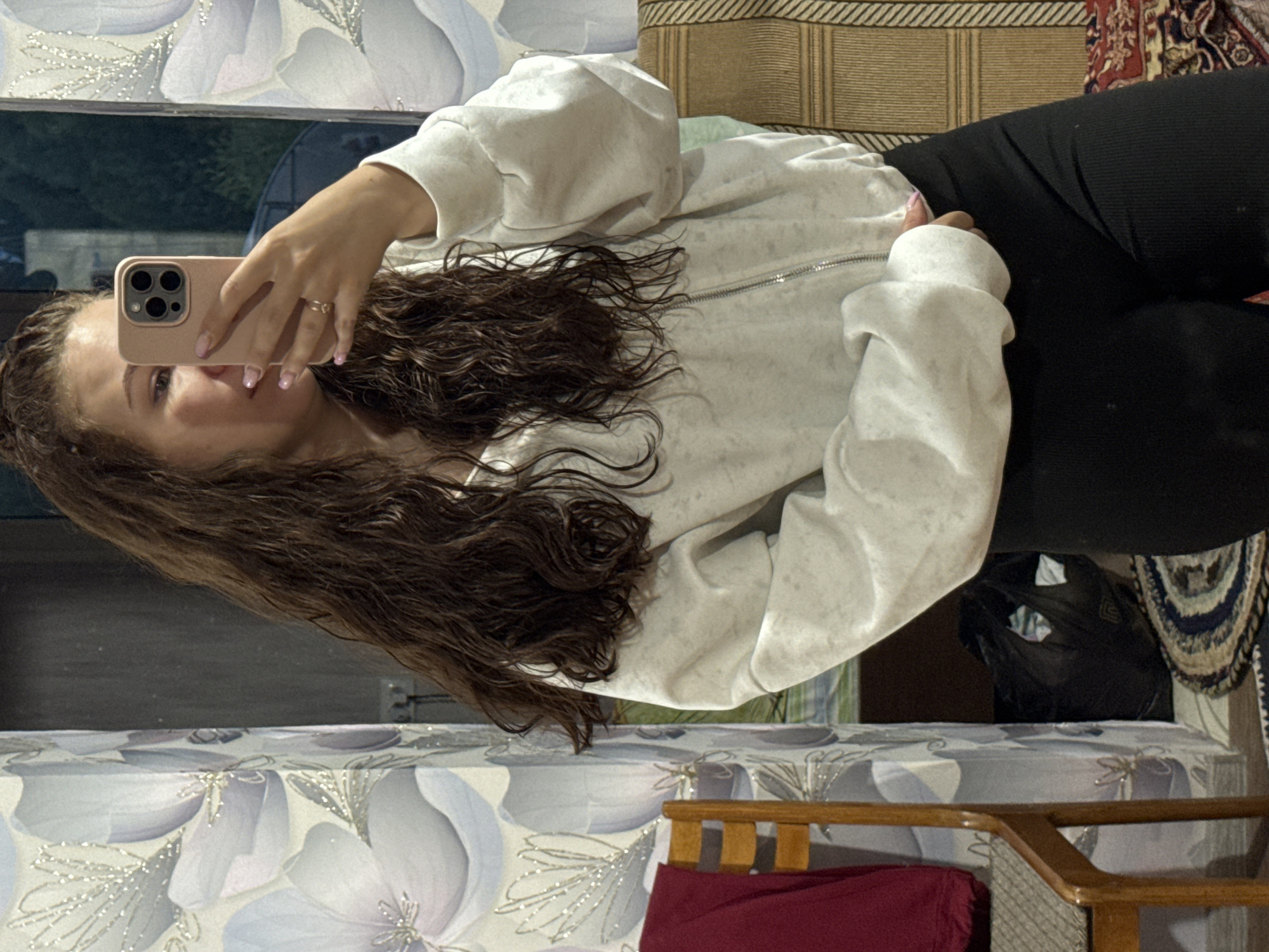 A young woman with long, brown, wavy hair is taking a selfie using a pink smartphone. She is wearing a light gray zippered sweatshirt and black leggings. She is wearing a gold ring. The background includes a floral pattern on what appears to be a bedspread or mattress, part of a wooden chair frame with red fabric, and a rug with various patterns. The location is not identifiable as the city of Nizhnyaya Gar', Russia from the given image.