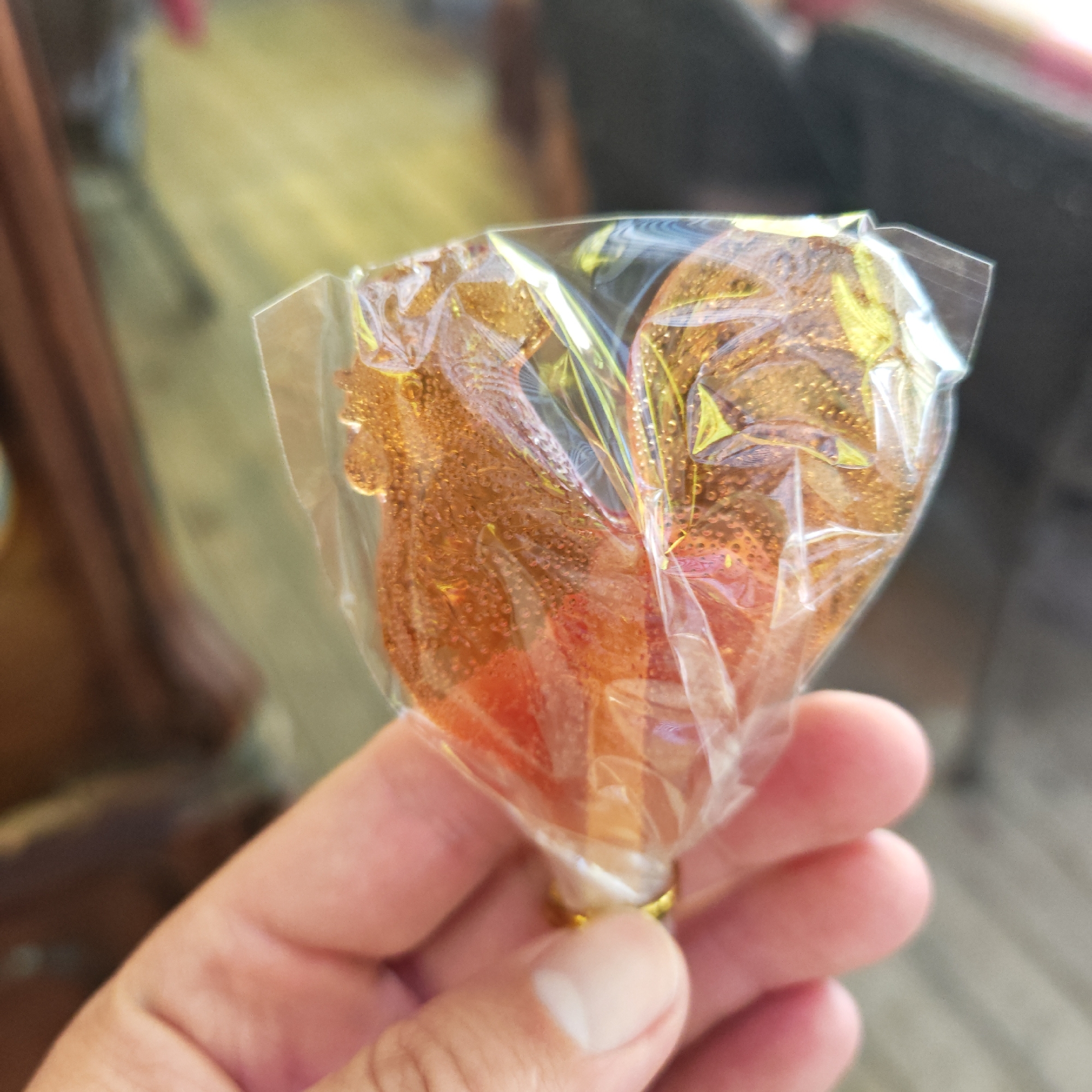A hand holds a rooster-shaped lollipop in clear plastic packaging. The lollipop appears to be amber or brown in color. A gold band is visible at the base of the lollipop where it meets the stick. The background shows an out-of-focus outdoor setting, possibly a patio or street scene. There is no confirmed information that the image was taken in Rostov, Russia.