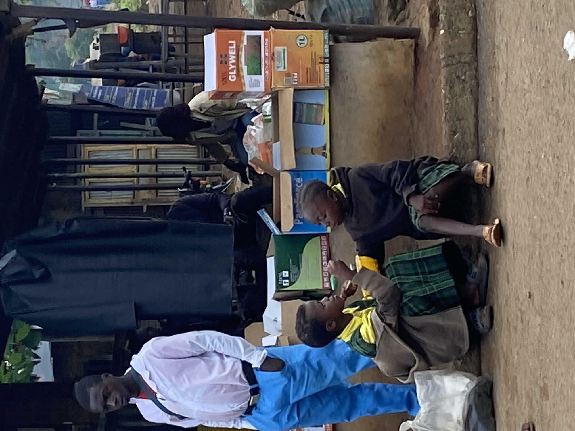 This image, set in Riyom, Nigeria, depicts an outdoor scene featuring several individuals and various objects, possibly in a street or market environment. The ground appears to be dirt or gravel.
In the foreground, a man stands dressed in a white long-sleeved shirt and blue trousers with a black belt, looking downwards. His expression is neutral. To his right, two young individuals are on the ground. One, wearing a yellow shirt and a grey jacket over a plaid skirt/kilt, is sitting or kneeling with their left hand raised towards the face of the other young person. The second young person, wearing a dark hoodie and green shirt, is kneeling facing the first, with their mouth slightly open, seemingly engaged in an interaction, perhaps eating or receiving something. One of these individuals is wearing open-toed sandals.
In the mid-ground, behind these individuals, several cardboard boxes are stacked. One prominent box displays the text "GLYWELI" and another indicates "1L" (likely for 1 liter). Another person, wearing dark clothing and a hat or head covering, is partially visible behind these boxes, appearing to be sorting items or working. A dark tarp or cover is stretched over a structure to the left. The background reveals a somewhat cluttered scene with metal bars, possibly part of a stall or fence, and distant foliage, suggesting an urban or semi-urban setting. The lighting indicates it is daytime, with soft, diffused light, suggesting an overcast day or shade. The overall atmosphere appears to be a candid slice of daily life.