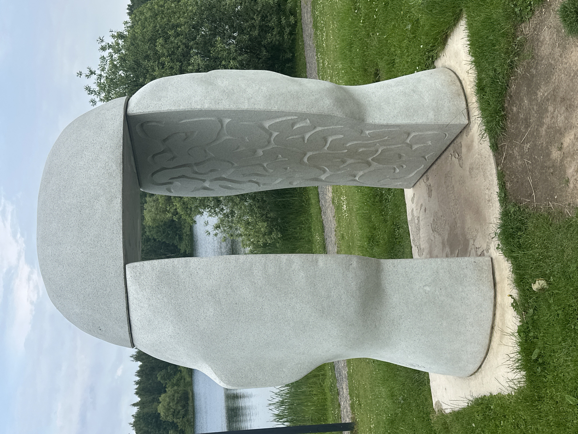An outdoor stone sculpture resembling an open head is situated on a concrete base in a park. The sculpture is divided into two halves, with the right half featuring carved patterns that suggest the texture of a brain. Behind the sculpture, a body of water, likely a lake, is visible, bordered by lush green trees and grass. The sky above is overcast with clouds. The sculpture is located in the Loshitsa Park in Minsk, Belarus.