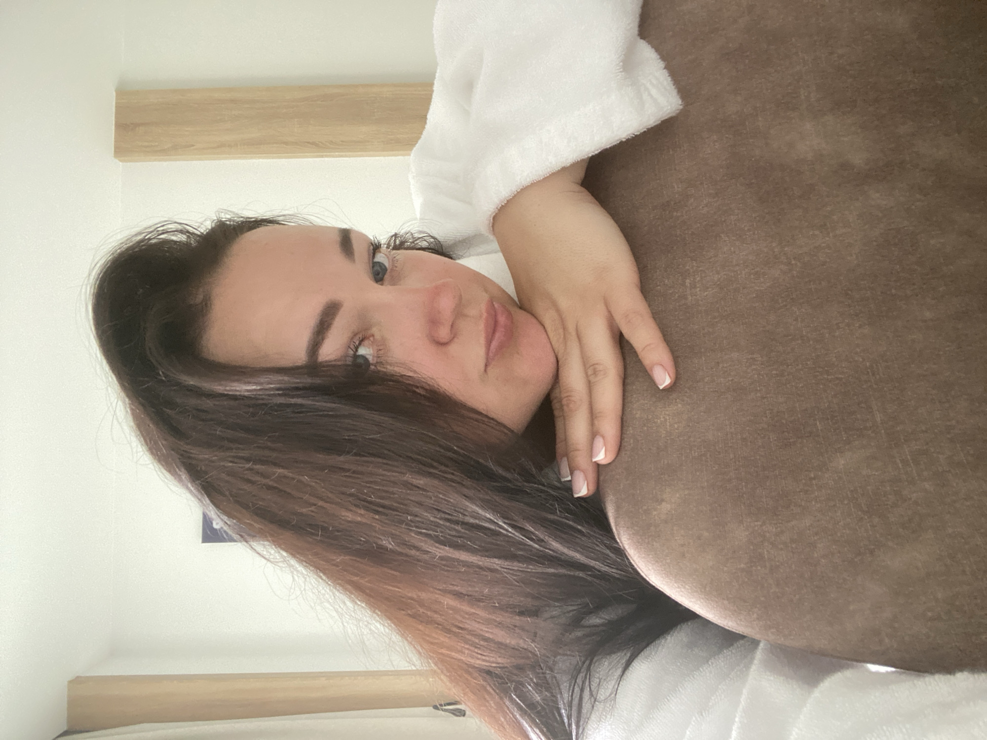 A person with dark brown hair and blue eyes is resting their head on a brown, textured surface while wearing a white robe. Their right hand is visible with manicured nails. The background includes a white wall and a light brown wooden shelf. No information within the image confirms this is in Sochi, Russia.