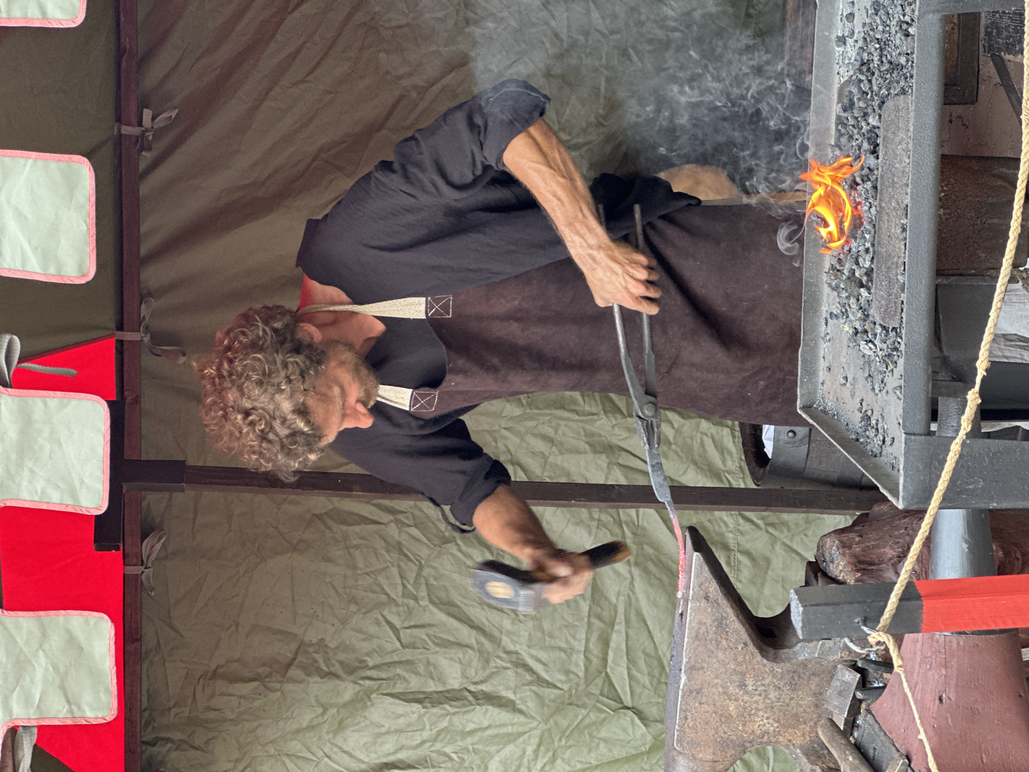 A man with curly blond hair and a beard, wearing a dark shirt and a brown apron, is blacksmithing in Vianden, Luxembourg. He is holding tongs with a glowing piece of metal in his right hand and a hammer in his left, poised to strike the metal on an anvil. A forge with a bright orange flame is visible in the foreground, emitting smoke. The blacksmithing area is set up under a tent with red and light green trim. The background consists of a wrinkled, olive-green tarp.