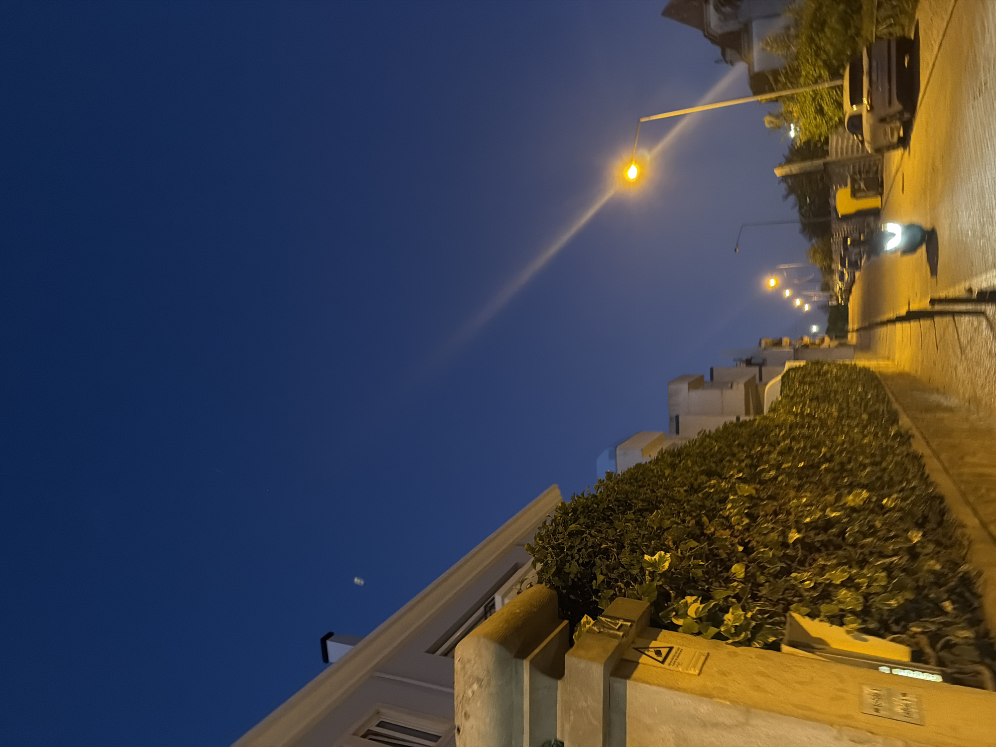 The image shows a street scene in Cascais, Portugal, at dusk or early evening. The sky is a deep blue, with a hint of a faint contrail or atmospheric phenomenon visible in the upper left quadrant, and a brighter, streaking light source diagonally bisecting the sky from the upper right to the center. On the left, a building with a white facade and a dark-capped chimney is partially visible, with a substantial hedge or ivy-covered wall running along its base. A yellow post with signage, including a triangular warning symbol, is positioned in front of the hedge.
The street itself is paved with cobblestones and slopes downwards from right to left. Several streetlights illuminate the scene, with one prominent streetlight in the mid-right casting a bright yellow glow and a long, diagonal beam across the sky. A white scooter with its headlights on is moving down the street towards the viewer. Further down the street, several cars are parked or moving, and more streetlights are visible. The overall atmosphere is tranquil and residential, characteristic of a European coastal town in the evening.