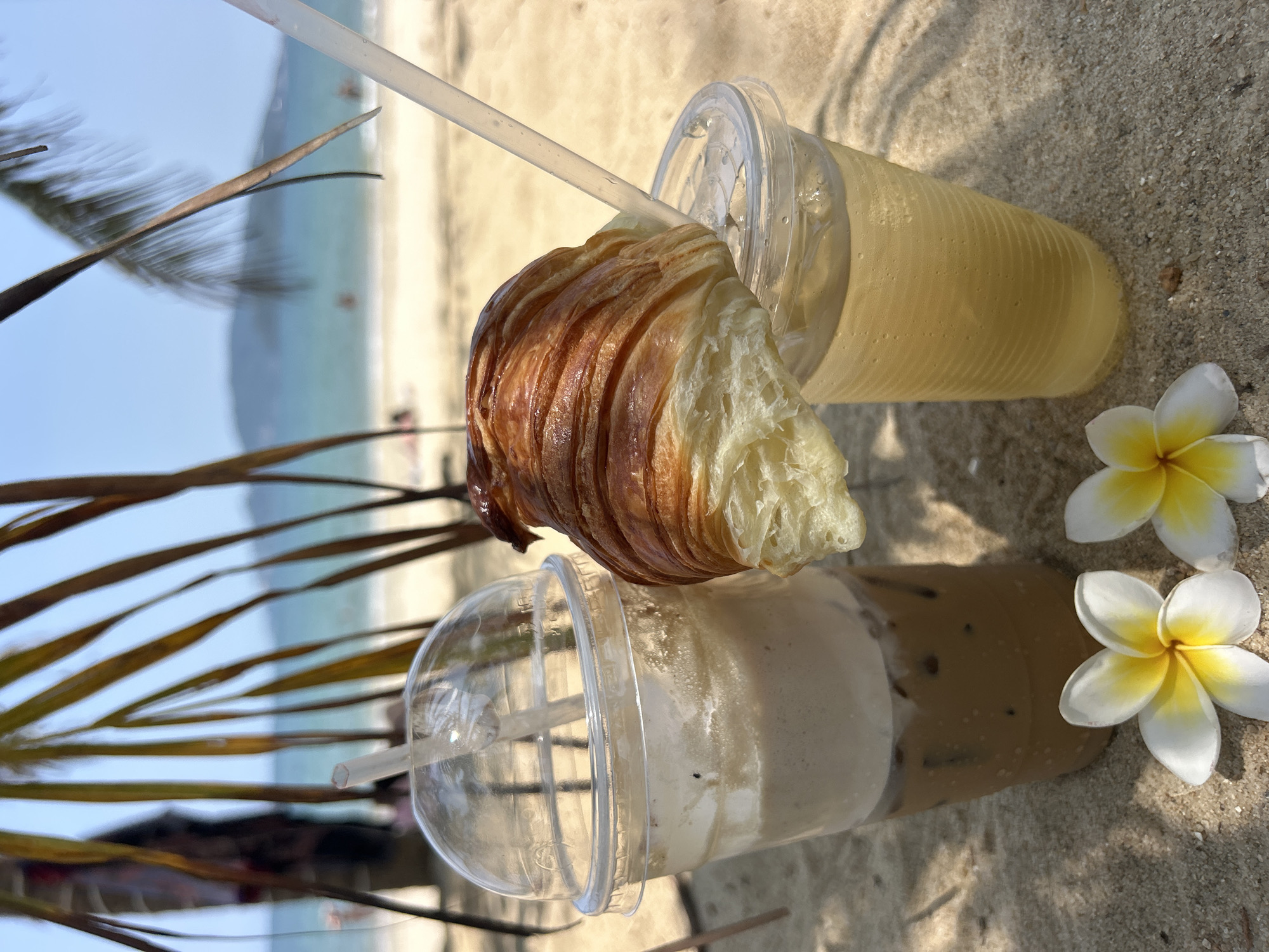The image presents a sun-drenched, tranquil beach scene featuring tropical refreshments. In the foreground, two clear plastic cups with drinks are placed on light-colored sand. The top cup contains a light yellow beverage, possibly juice, with a clear straw. A golden-brown, flaky croissant, partially broken to expose its soft interior, rests on the rim of this cup. Below it, the second cup holds an iced coffee, displaying distinct layers of creamy foam and dark liquid, also equipped with a straw.
Beside the lower cup, two vibrant white and yellow plumeria flowers add a decorative touch to the setting. Palm fronds extend into the frame from the left, casting dappled shadows on the sand, indicating bright daylight, likely mid-morning or early afternoon.
The background reveals a serene coastal landscape. A calm, light blue ocean stretches into the distance, meeting a faint outline of hills or mountains on the horizon. Small, indistinct figures are visible on the beach and in the water, suggesting a peaceful yet active beach environment. The overall impression is one of relaxation and leisure in a beautiful tropical location, consistent with a setting like Nha Trang, Vietnam. No text is visible in the image.