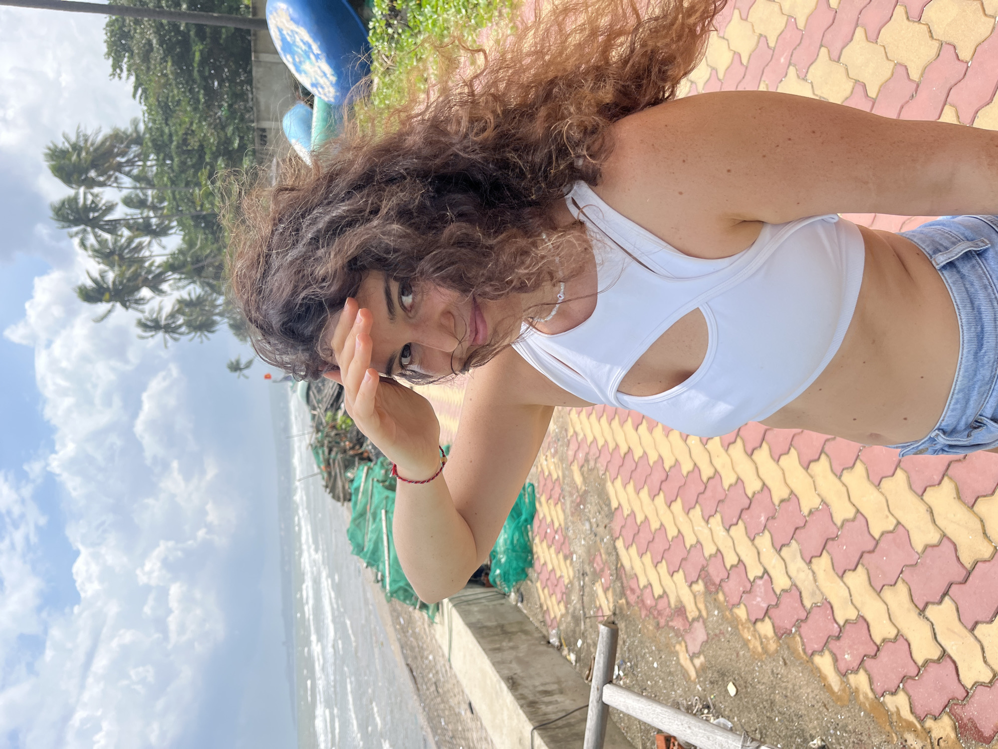 In Mũi Né, Vietnam, a young woman with long, curly brown hair stands on a paved walkway near the beach. She is wearing a white cutout crop top and blue denim shorts. Her right hand is raised to shield her eyes as she looks towards the ocean, which is visible in the background. The beach is lined with palm trees and some fishing nets are piled on the shore. The sky is cloudy with patches of blue. The walkway is paved with interlocking blocks of red and yellow.
