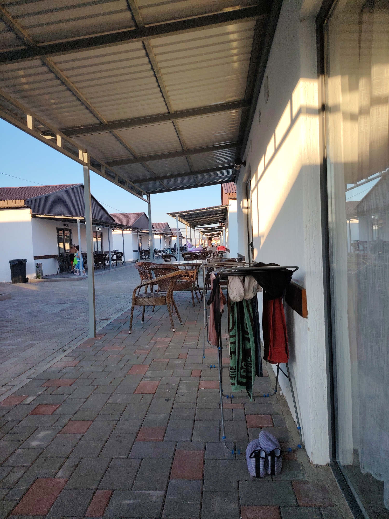 The image depicts an outdoor walkway next to a white building with a corrugated metal roof extension, likely in the city of Toranghylyk, Kazakhstan. The walkway is paved with rectangular red and gray interlocking bricks. Several tables and chairs made of woven material are set up under the roof extension, some with umbrellas. A clothes drying rack with several towels and garments hanging on it is positioned against the building wall. A pair of slippers rests on the ground near the drying rack. In the background, people are visible walking and sitting at tables, and more buildings with similar roof extensions are seen lining the street. The sun casts long shadows on the wall and ground, indicating it is late afternoon or early evening.
