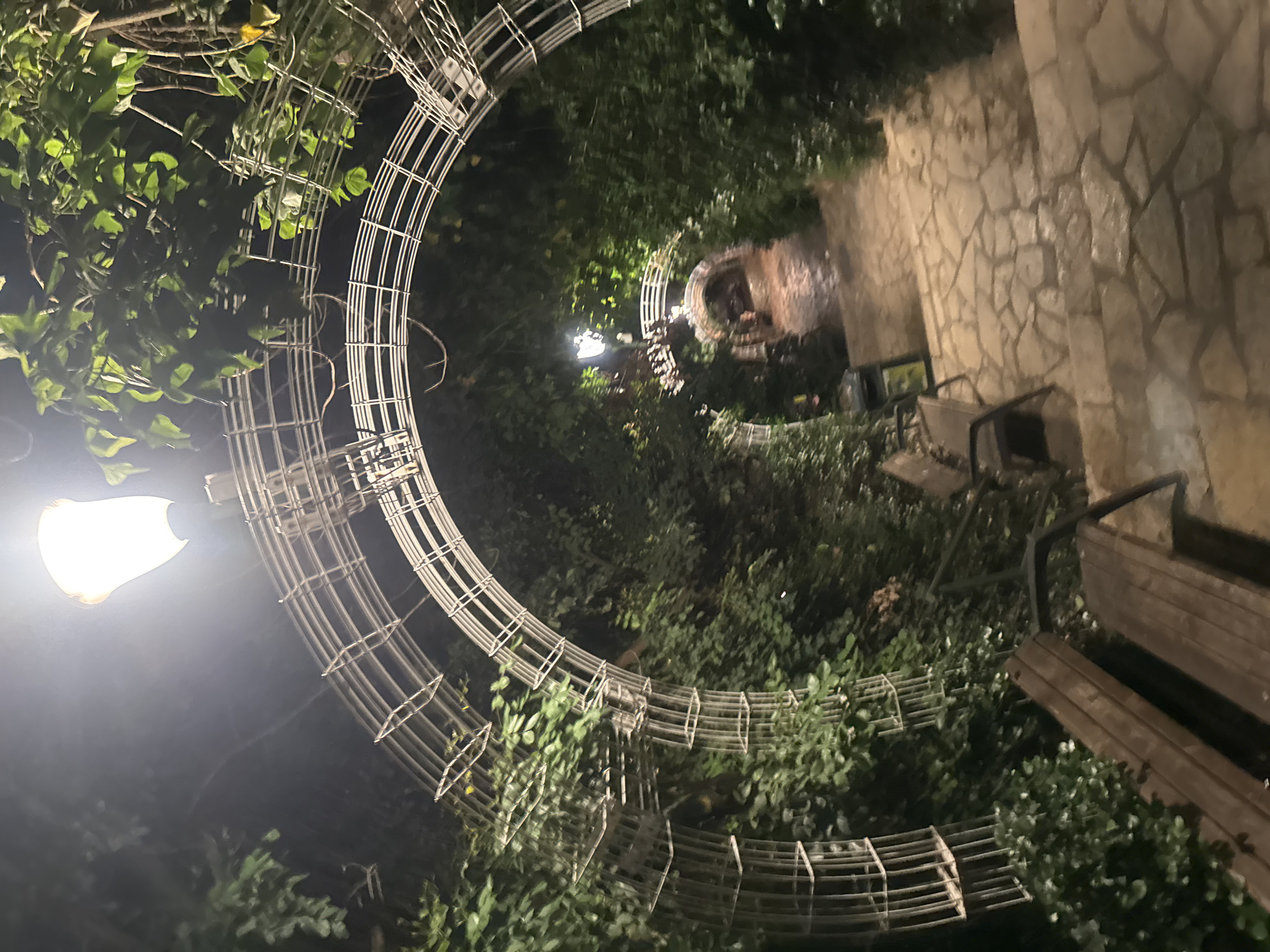 Captured at night in Ramat Gan, Israel, the scene presents a pathway illuminated by a lamppost and a series of arched metal structures. Green foliage is interspersed with the structures, adding depth and texture. The pathway leads towards a stone wall with benches adjacent to it. The composition creates a sense of depth and enclosure.