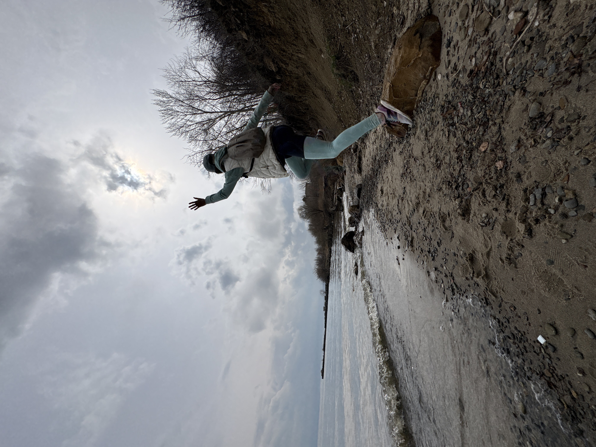 A woman stands at the edge of a sandy beach bordering a body of water, likely the Volga River, at Ulyanovsk, Russia. The sky is overcast with gray clouds. The woman, wearing light blue leggings, a light-colored jacket, and sneakers, is positioned with one foot on the sand and one foot on a rock, seemingly balancing with her arms outstretched. The shoreline consists of sand and pebbles, with some vegetation and bare trees visible along the bank. The water is choppy, and the overall scene suggests a cool, overcast day.