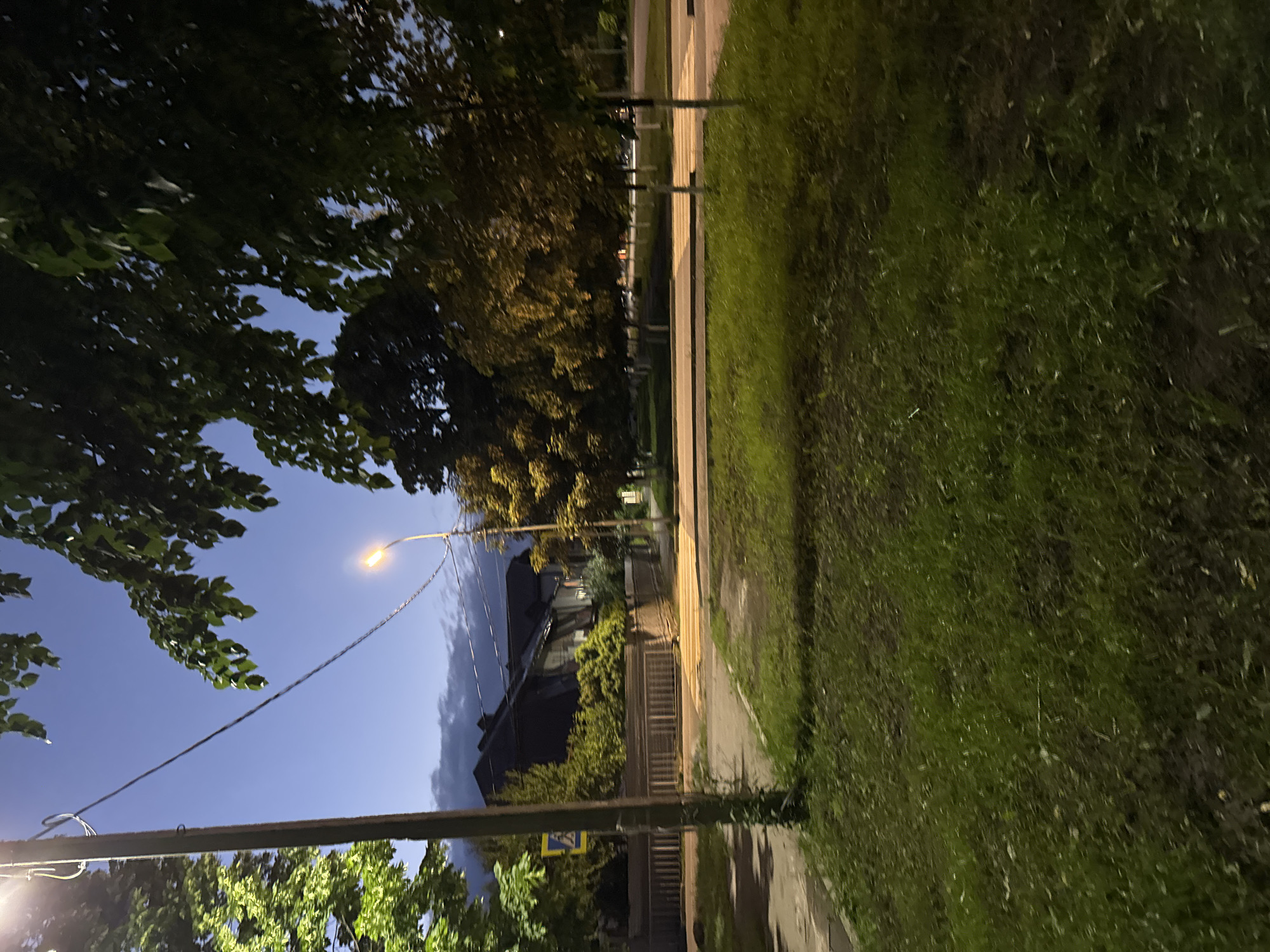 The image depicts a street scene at dusk or night in Karachayevsk, Russia. In the foreground, there is a grassy area. A wooden utility pole stands prominently to the left, with wires extending across the frame. Beyond the pole is a sidewalk and a road with a pedestrian crossing. A fence runs along the property line of a building complex visible in the middle ground. The buildings have dark roofs and appear to be residential or administrative. Trees with green and yellow foliage line the street, some illuminated by streetlights. The sky is a deep blue with some cloud cover. A street sign is visible on the left side of the image.