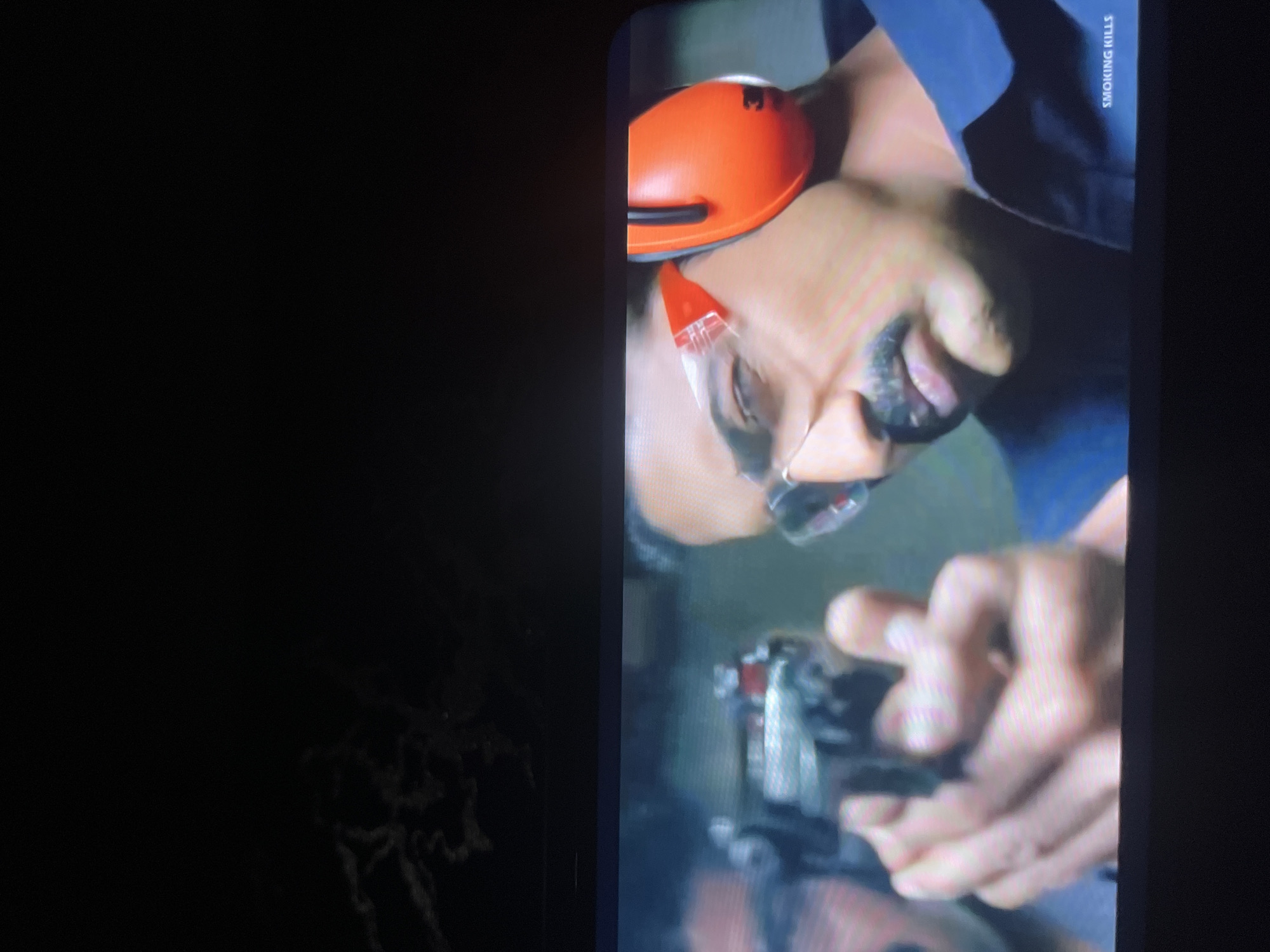 The image shows a close-up of a man aiming a handgun. He is wearing protective orange earmuffs and clear safety glasses. His face is partially obscured by his hand holding the gun. The background is blurred, but appears to be an indoor setting, possibly a shooting range. There is text in the bottom right corner that reads "SMOKING KILLS". There is no indication of the location being in Bukuru, Nigeria or any other specific city.