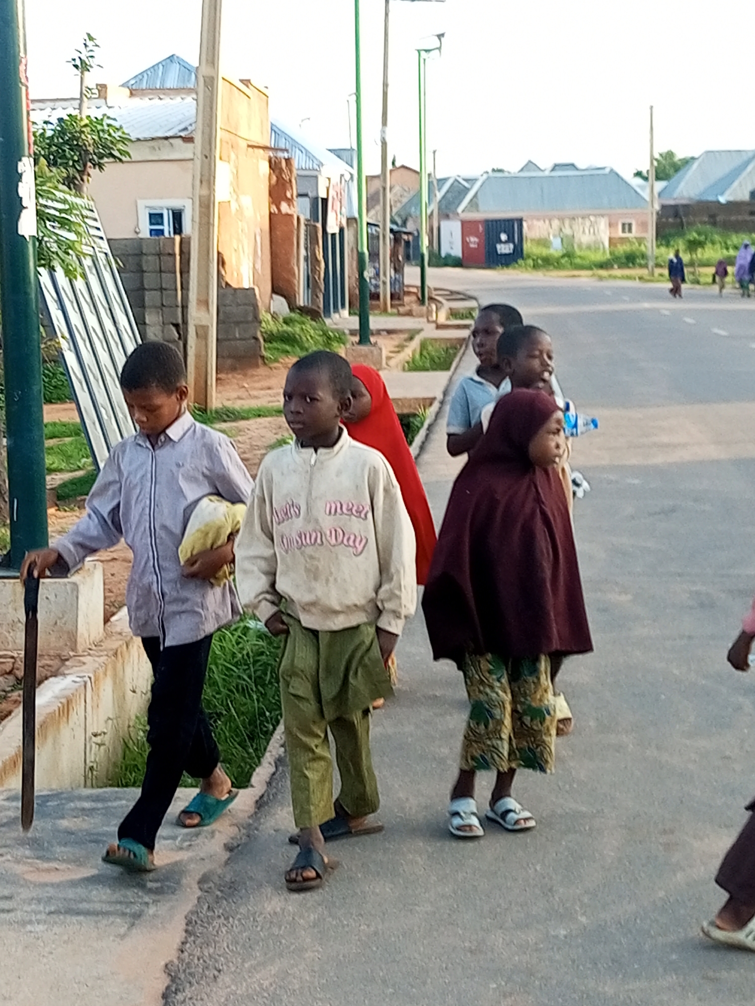 In Tempure, Nigeria, several children walk along a paved road. In the foreground, a young boy in a light purple button-down shirt and black pants, holding a yellow cloth, walks to the left. To his right, another boy wears a cream-colored sweatshirt with text and green pants. A third child, wearing a dark maroon hijab and patterned pants, walks to the right of the second boy. In the background, other children and adults are visible. The environment includes buildings with corrugated metal roofs, a streetlamp, and vegetation.