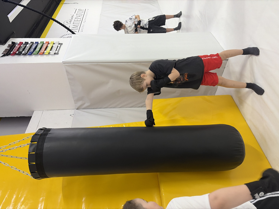 The image displays an indoor martial arts training facility, identified by the signage "Император Школа Единоборств" (Emperor School of Martial Arts), located in Novosaratovka, Russia. The facility features yellow and white padded walls and a light-colored floor.
A black cylindrical punching bag, suspended by chains, hangs against a yellow padded wall. In the center, a young male child with light brown hair, wearing a black t-shirt, red shorts, and black fingerless gloves, is seen striking the punching bag with his right hand. His left hand is in a guard position, and his focus is on the bag.
To the immediate left, partially visible, is another young male facing the punching bag, wearing a white t-shirt and dark shorts. In the background to the right, a third young male with dark curly hair, dressed in a patterned white shirt, dark shorts, and dark socks, is standing and adjusting a black belt around his waist. Mounted on a white wall section in the background is a display of colored martial arts belts, labeled with Russian text including "JUDO" and various principles such as "ВЕРНОСТЬ" (Loyalty) and "ЧЕСТНОСТЬ" (Honesty). A thick black rope also hangs from the ceiling in the background.