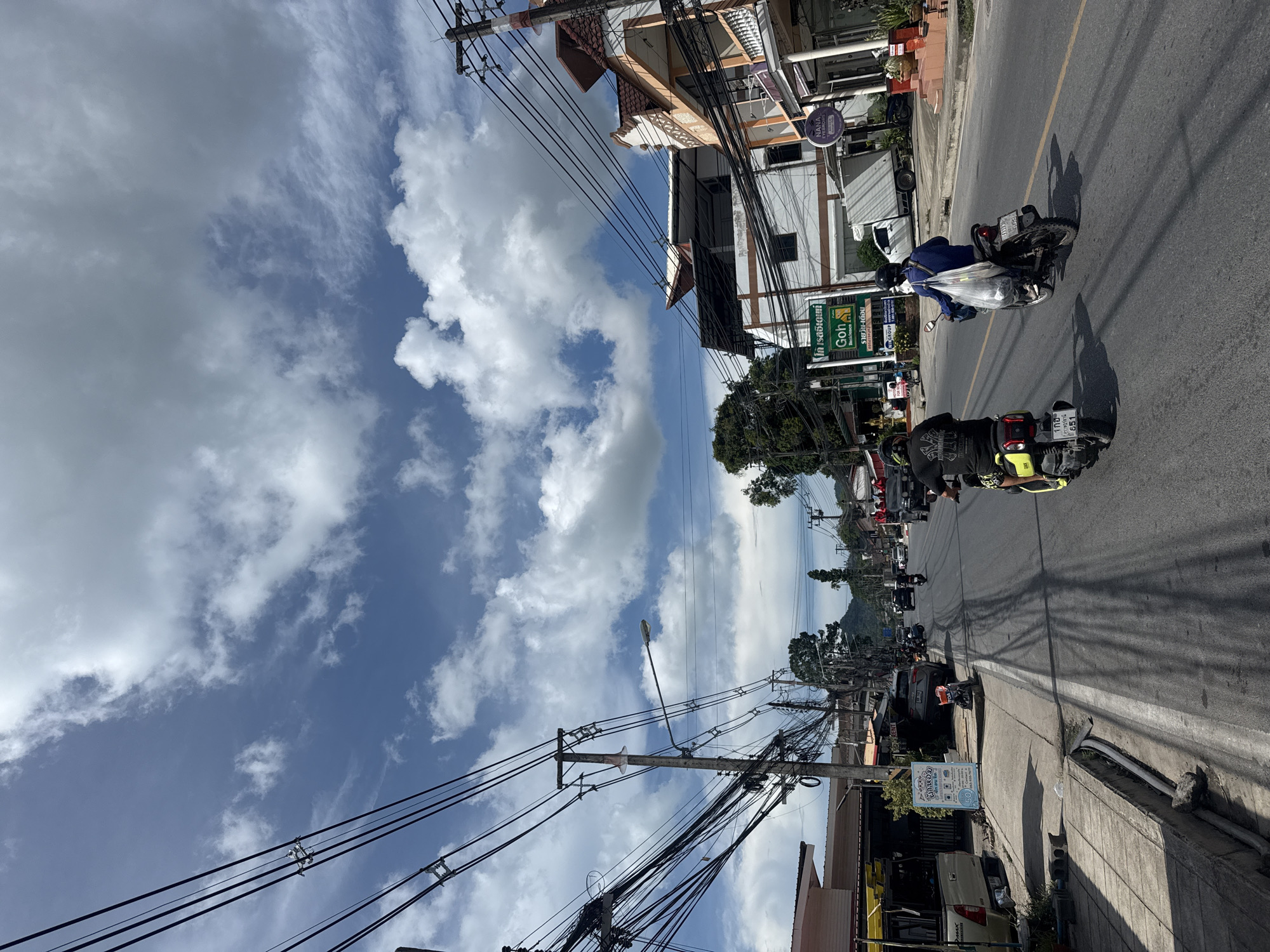 The image depicts a street scene in Baan Chaweng Noi, Thailand. The day is bright with a partly cloudy sky. The road is paved and lined with buildings and utility poles. There are several motorcycles on the road, with two riders clearly visible. One rider is on a yellow and black motorcycle, wearing a black shirt with yellow accents and a black helmet. Another rider on a black motorcycle is wearing a blue jacket and carrying a bag. Several cars are also present on the road, along with a delivery truck in the background. Buildings on either side of the street appear to be shops and residences. Signs in Thai script are visible on some of the buildings. The scene suggests a busy commercial and residential area.