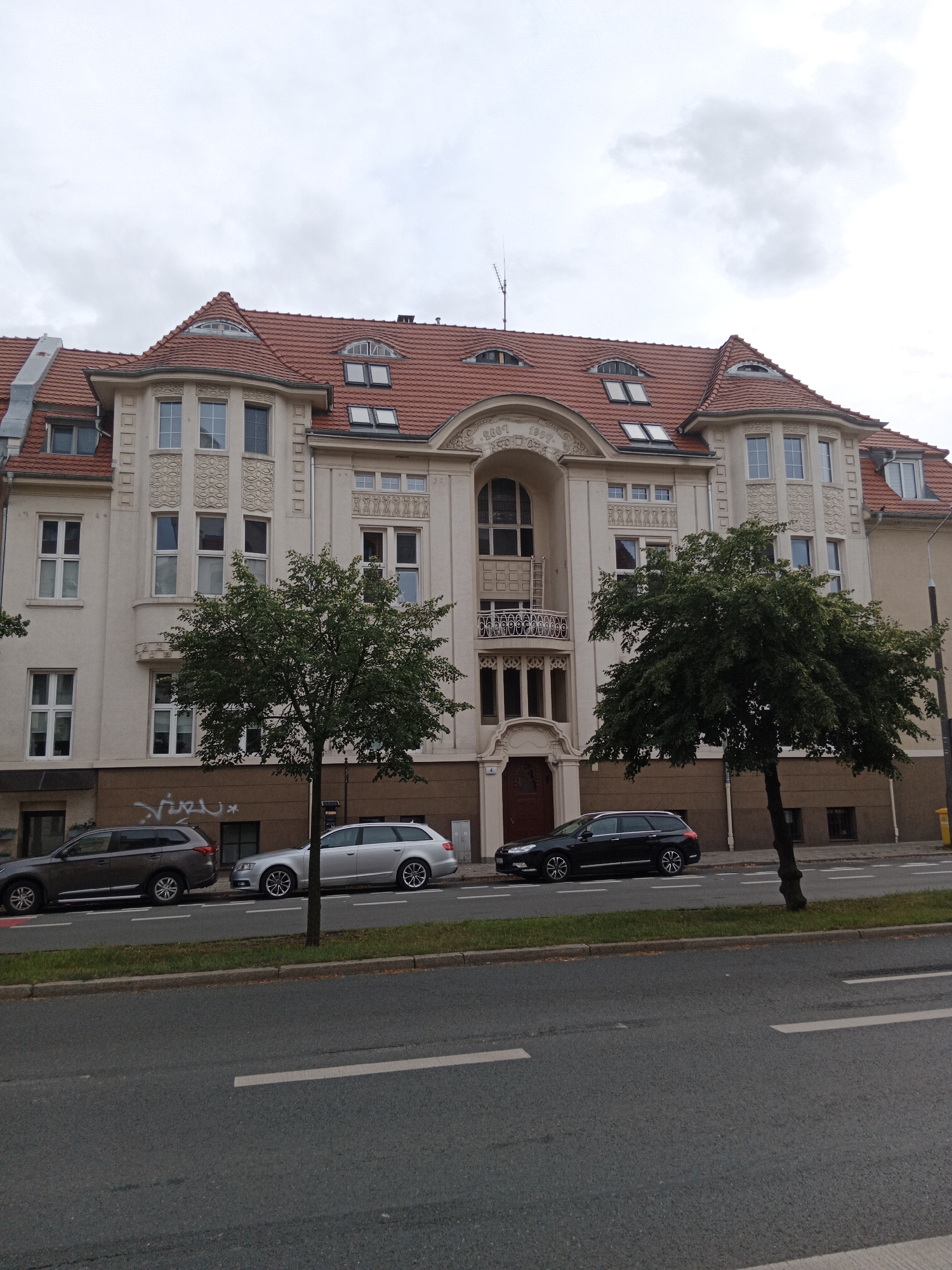 The image shows a street scene in Bydgoszcz, Poland, featuring an ornate apartment building with a red tiled roof and several cars parked along the sidewalk. The building, likely dating from the early 20th century, exhibits classical architectural elements with detailed ornamentation around the windows and a prominent arched entrance. The facade is a light cream color, and the roof has several dormer windows. Three cars are visible: a dark gray SUV on the left, a silver station wagon in the middle, and a black station wagon on the right. Two green trees are planted between the sidewalk and the street. The street itself is asphalt with white lane markings. The sky is overcast, suggesting a cloudy day. There are no people visible in the image. The overall impression is of a well-maintained urban street.