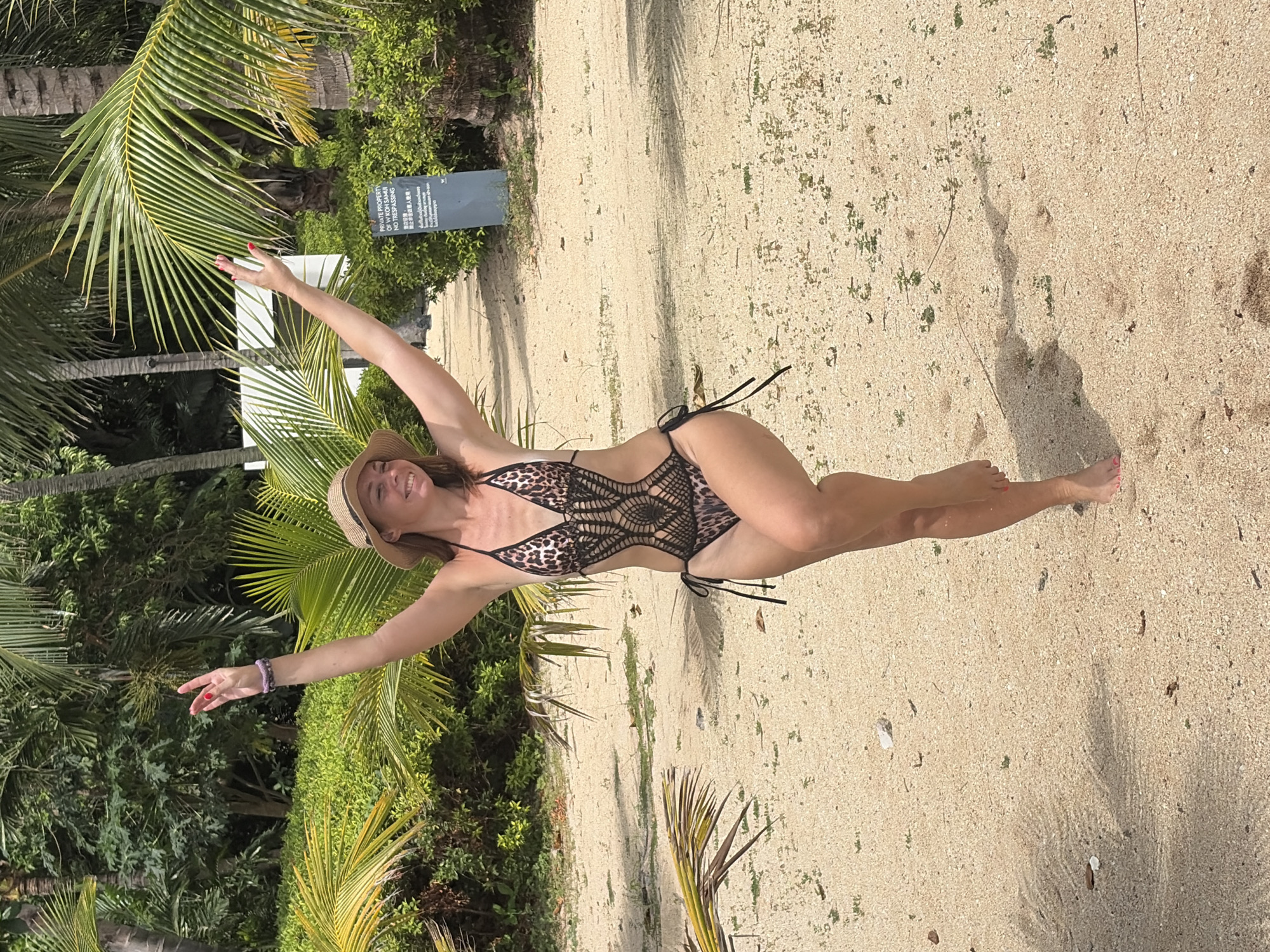 A woman is standing on a sandy beach, with her arms raised in the air. She is wearing a tan straw hat and a leopard-print one-piece swimsuit with black crochet detailing. Her left leg is bent, and her right leg is extended, with her right foot lifted slightly off the sand. She is smiling and looking to her right. The background consists of lush green foliage, palm trees, and a pathway. The sand on the beach is light brown and speckled with small green plants and some debris. A sign is visible in the background to the right, partially obscured by a palm frond, with text that appears to be in English and another language. The lighting suggests it is daytime, with some dappled shade present. The location is identified as Baan Bo Phut, Thailand.