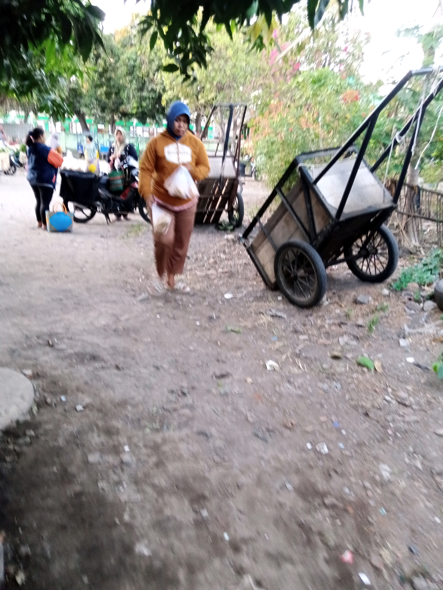 In Sono Ageng, Indonesia, a woman wearing a blue hijab and a mustard-colored hoodie, holding a plastic bag, walks on a dirt path. She is facing towards the camera and her legs are visible from the knees down, clad in brown pants. To her left, a motorcycle is parked, and another woman in a dark jacket and blue pants stands beside it. In the background, trees with green foliage and some blooming pink flowers line the area, with other people and carts visible in the distance. To the right, a black wheeled cart with a wooden base stands on the dirt. The ground is a mixture of dirt, small stones, and scattered debris.