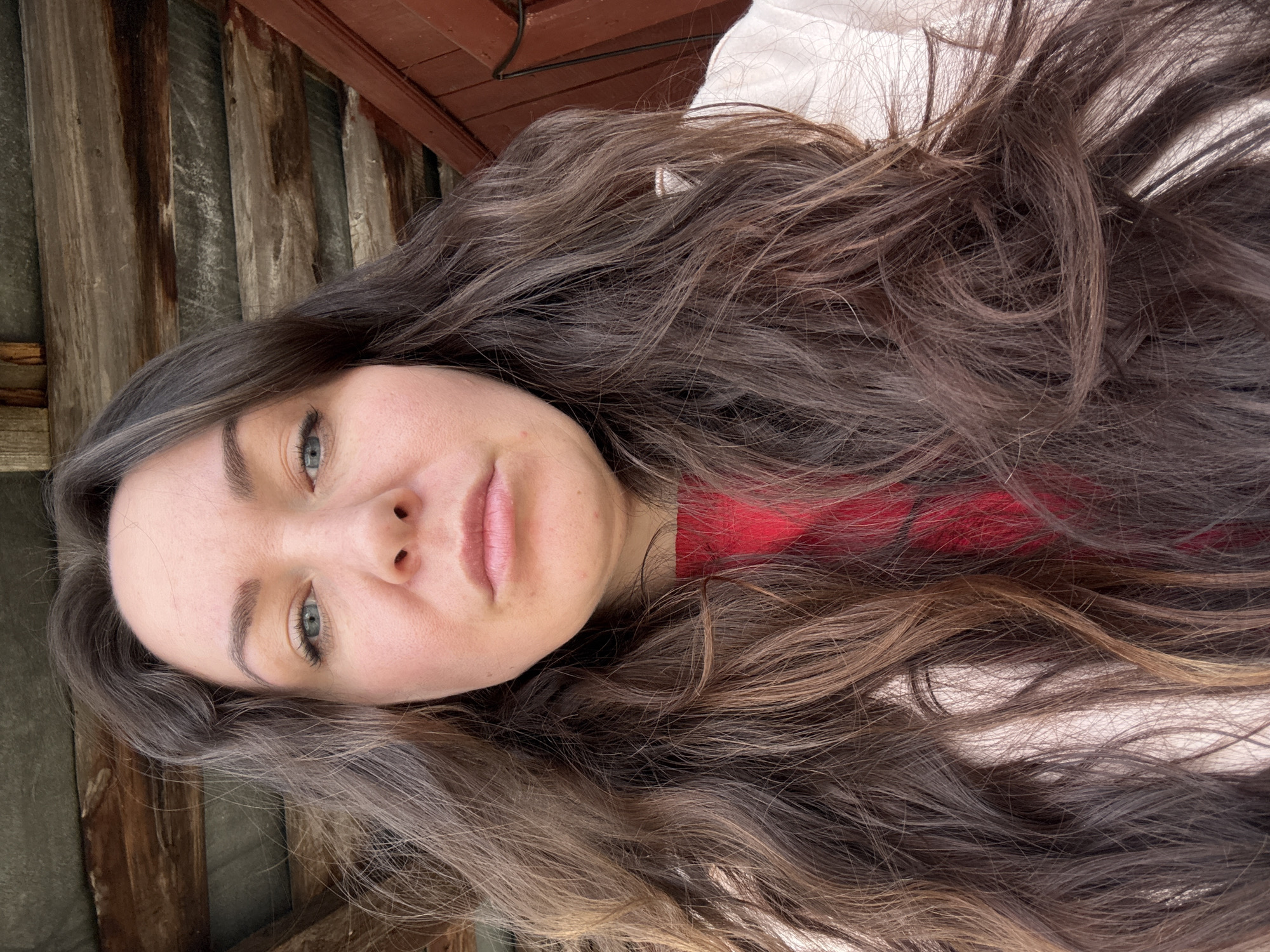 A selfie of a woman with long, wavy brown hair. She has fair skin, light blue eyes, and light pink lips. She's wearing a red shirt and a white top, possibly a jacket or blouse. The background consists of wooden planks. There is no indication this image was taken in the city of Chaikovsky, Russia.