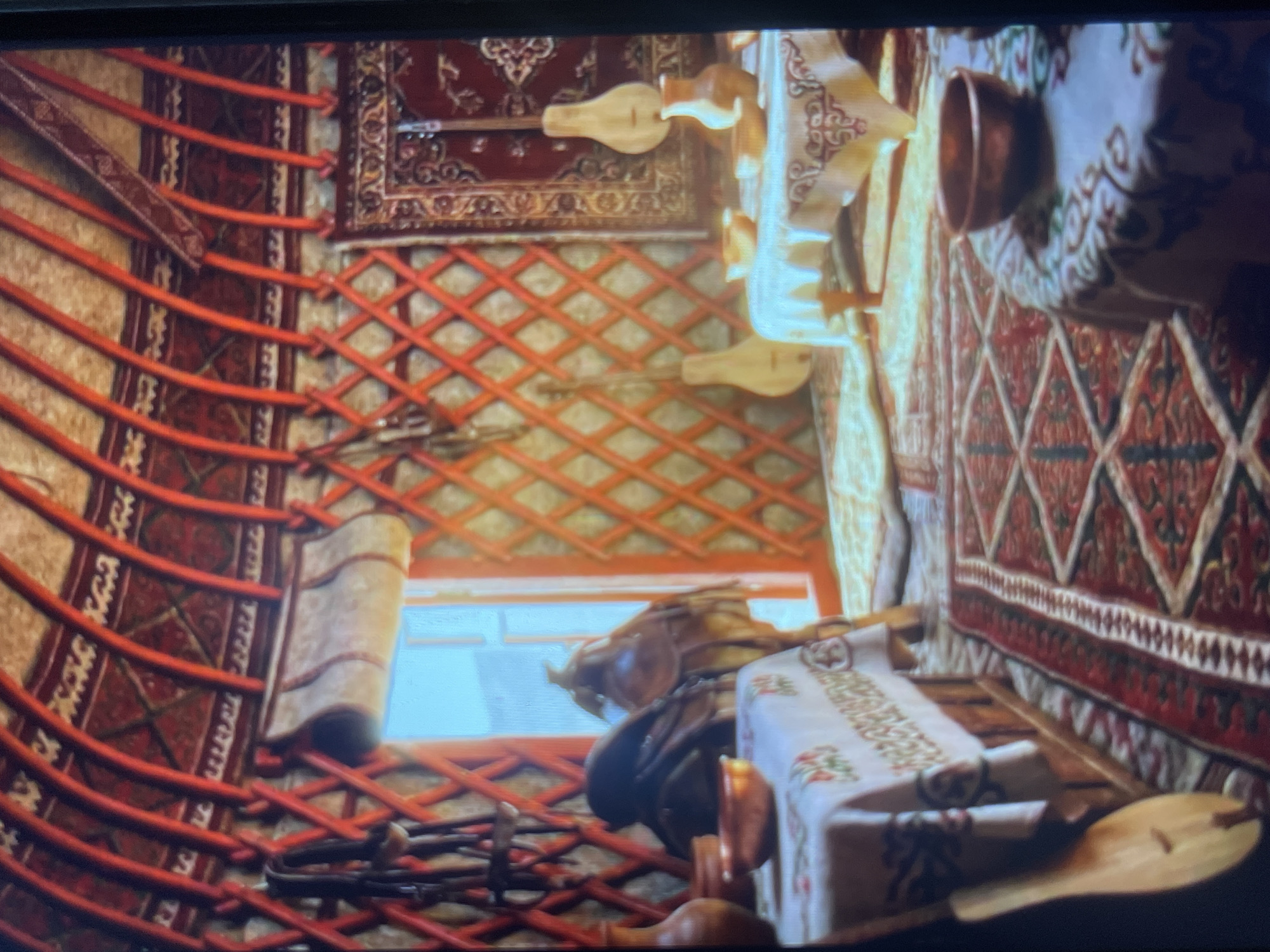 The image shows the interior of a yurt, a traditional nomadic dwelling. The yurt's structure is visible, with curved wooden poles forming the roof and a lattice of wooden supports forming the walls. These structures are draped with rich, patterned textiles in red, white, and gold. Several musical instruments, likely dombras, with their distinctive pear-shaped bodies and long necks, are leaning against the walls. Two low tables covered with patterned cloths are set with copper bowls, suggesting a gathering or meal. A saddle and bridle hang on the wall near a window, and a large rug with a complex geometric pattern covers the floor. The overall impression is one of a well-appointed and culturally rich living space. The image does not provide specific details that would allow for pinpointing its location within Bishkek City, Kyrgyzstan.