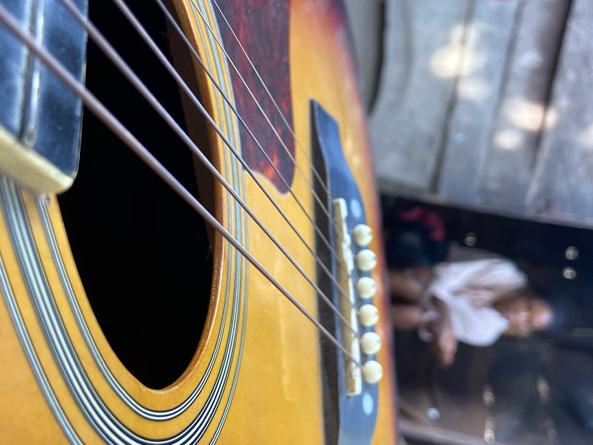 A close-up shot of an acoustic guitar with six strings dominates the foreground, showcasing the instrument's soundhole, pickguard, and bridge. The guitar's body features a warm, amber hue, contrasting with the dark interior of the soundhole. The strings are taut and appear to be made of metal.
In the blurry background, a person is discernible. The location of this scene cannot be verified within the city of Mararaba, Nigeria.