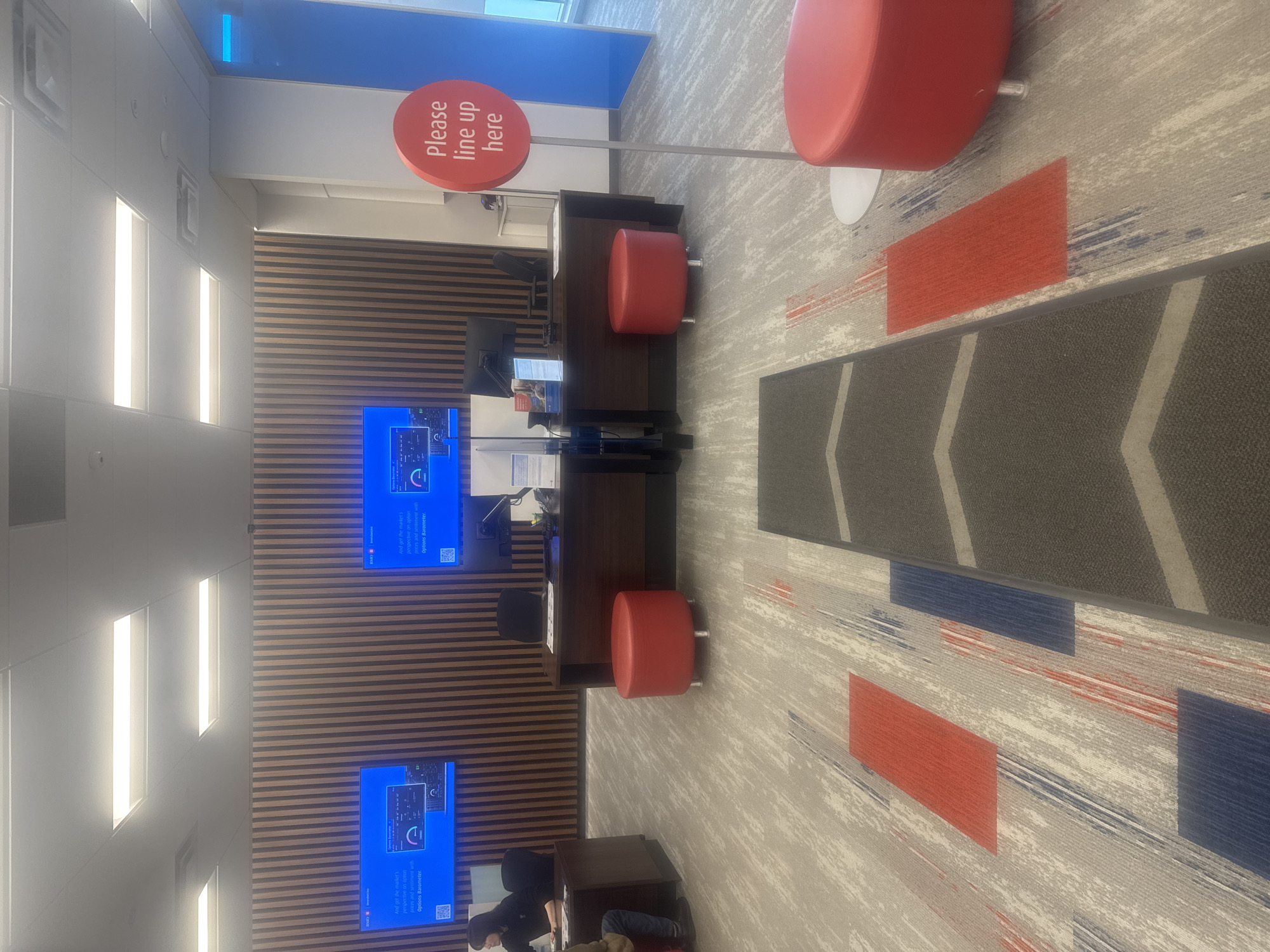 The image depicts the interior of what appears to be a bank or financial institution, likely located in Burnaby, Canada. The scene features a modern design with a light-colored carpeted floor adorned with abstract geometric patterns in red, blue, and gray. A row of vertical wooden panels forms a decorative backdrop for two large, wall-mounted monitors displaying financial data. In front of the monitors are desks with computers and office supplies. To the right, a circular red sign on a silver pole reads "Please line up here." Two red cylindrical ottomans are placed in the open area. The lighting is provided by fluorescent ceiling panels. The overall impression is one of a clean, organized, and contemporary service environment.