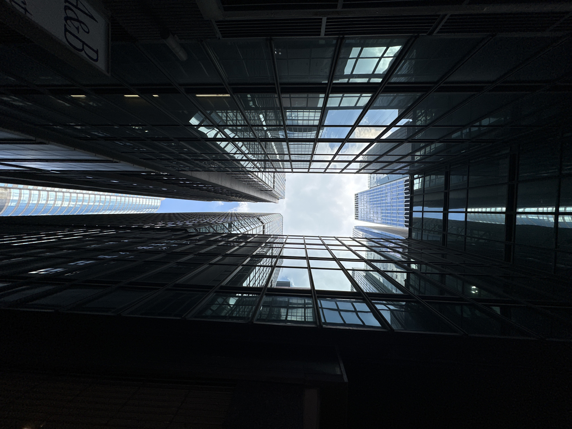 This is an extreme low-angle, upward shot of modern skyscrapers in a dense urban environment, likely Hong Kong, China, given the architectural style and building density. The perspective emphasizes the height and sheer glass facades of the buildings, creating a sense of being dwarfed by the structures.
The buildings are tall, sleek, and primarily made of glass and dark metal. Their reflective surfaces mirror the sky and other surrounding buildings, creating a complex geometric pattern. The sky visible between the buildings is a mix of white clouds and blue.
On the right side of the frame, a portion of a sign with the letters "d&b" is visible, suggesting a commercial establishment. The overall impression is one of a bustling, modern cityscape, with a focus on architectural design and the overwhelming scale of urban development. There are no people or animals visible in the image. The lighting suggests it is daytime, with the sun casting reflections on the buildings.