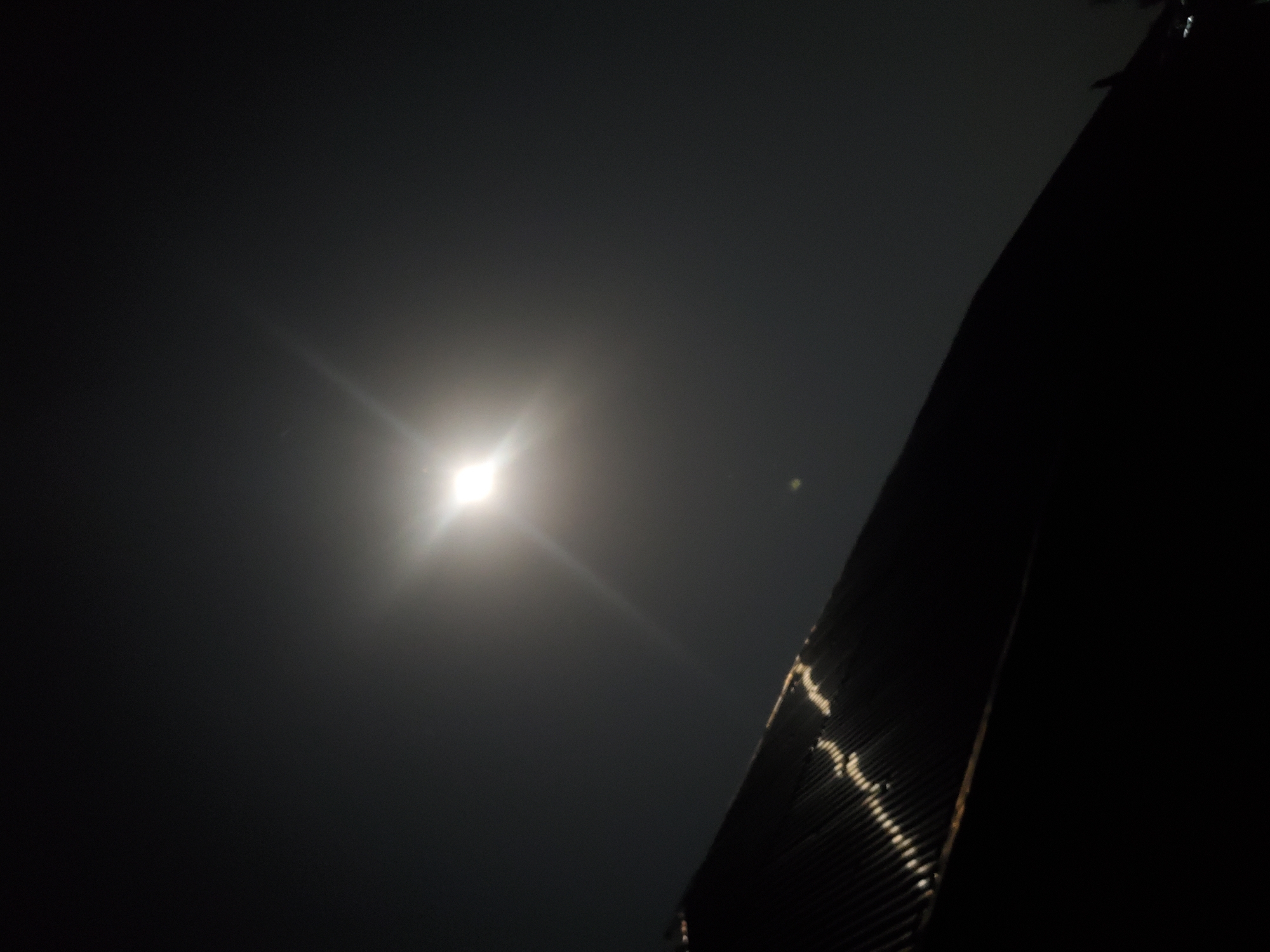 The image shows a night sky with a bright, starburst-like light source, likely the moon or a strong artificial light, positioned in the upper center. Below this, a dark, angled structure, appearing to be a corrugated metal roof, extends from the left side of the frame towards the right. The roof reflects some light, showing a pattern of highlights. The overall scene is dark, with the sky filling most of the frame, suggesting an outdoor setting, possibly in Kason, Nigeria, at night.