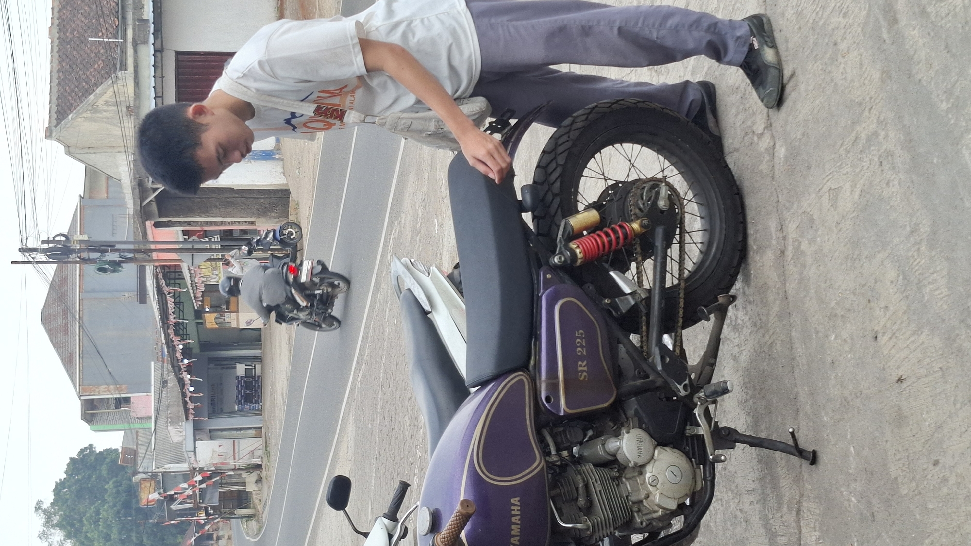 A young East Asian male wearing a white t-shirt and grey pants stands next to a purple Yamaha SR 225 motorcycle on a paved road in the city of Cisalak, Indonesia. He is looking down at the motorcycle, with his right hand near the seat. The motorcycle is parked on a concrete surface. In the background, there are buildings, trees, and another motorcycle with a rider. The sky is overcast. The scene suggests a roadside setting in a town or city.