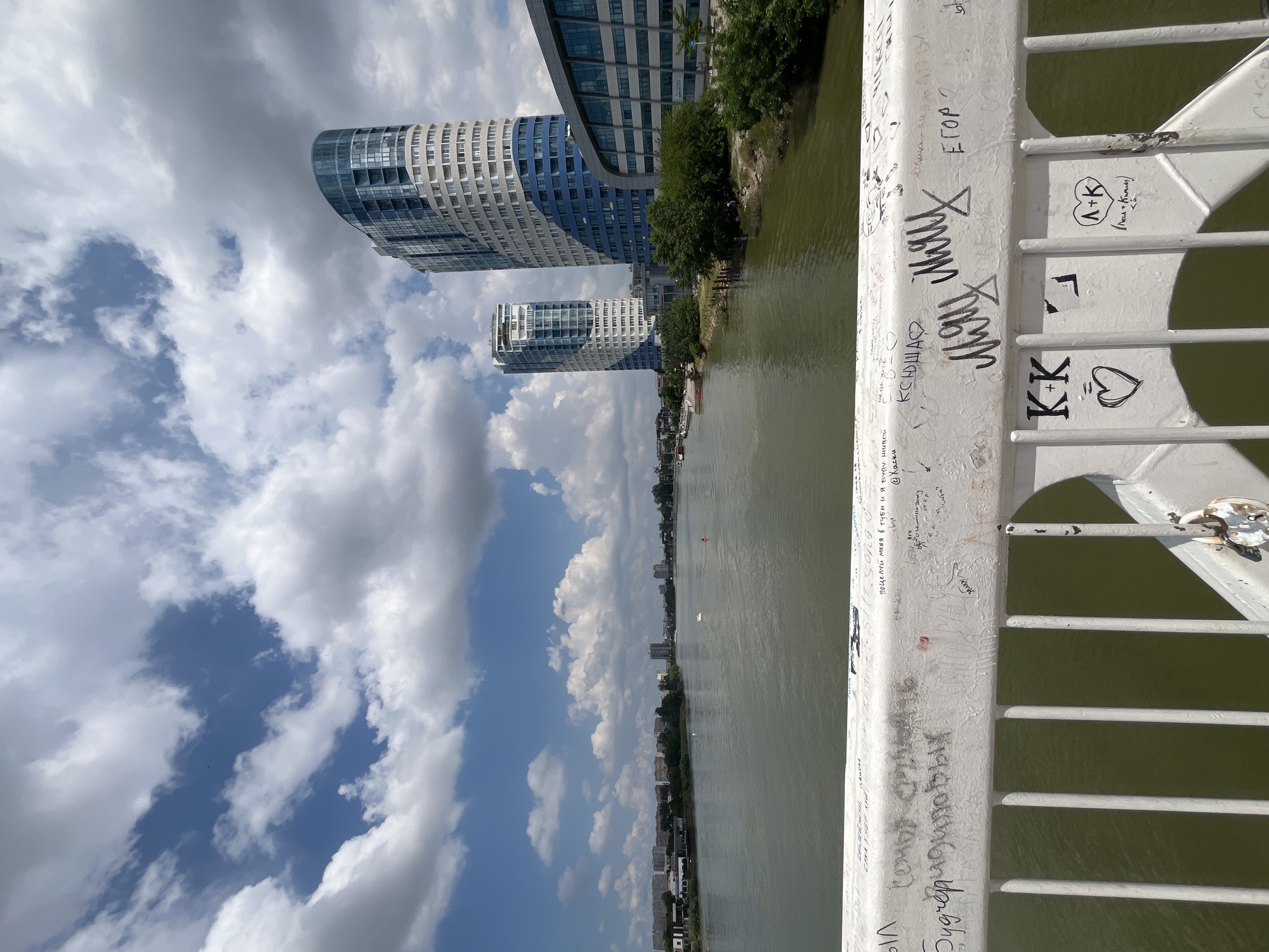 A wide shot shows the Krasnodar Reservoir with the Kuban River flowing through the city of Krasnodar, Russia. The water is a murky green, reflecting a partly cloudy sky. In the background, a city skyline is visible with apartment buildings and trees. In the foreground, a white railing is covered with graffiti, including names and symbols. The railing is a part of a bridge, with vertical white bars extending down. Several modern, curved residential buildings with blue and white facades stand on the right bank of the river, dominating the middle ground. One building has a semicircular design, and another is taller and more cylindrical. The overall impression is an urban landscape with elements of nature.