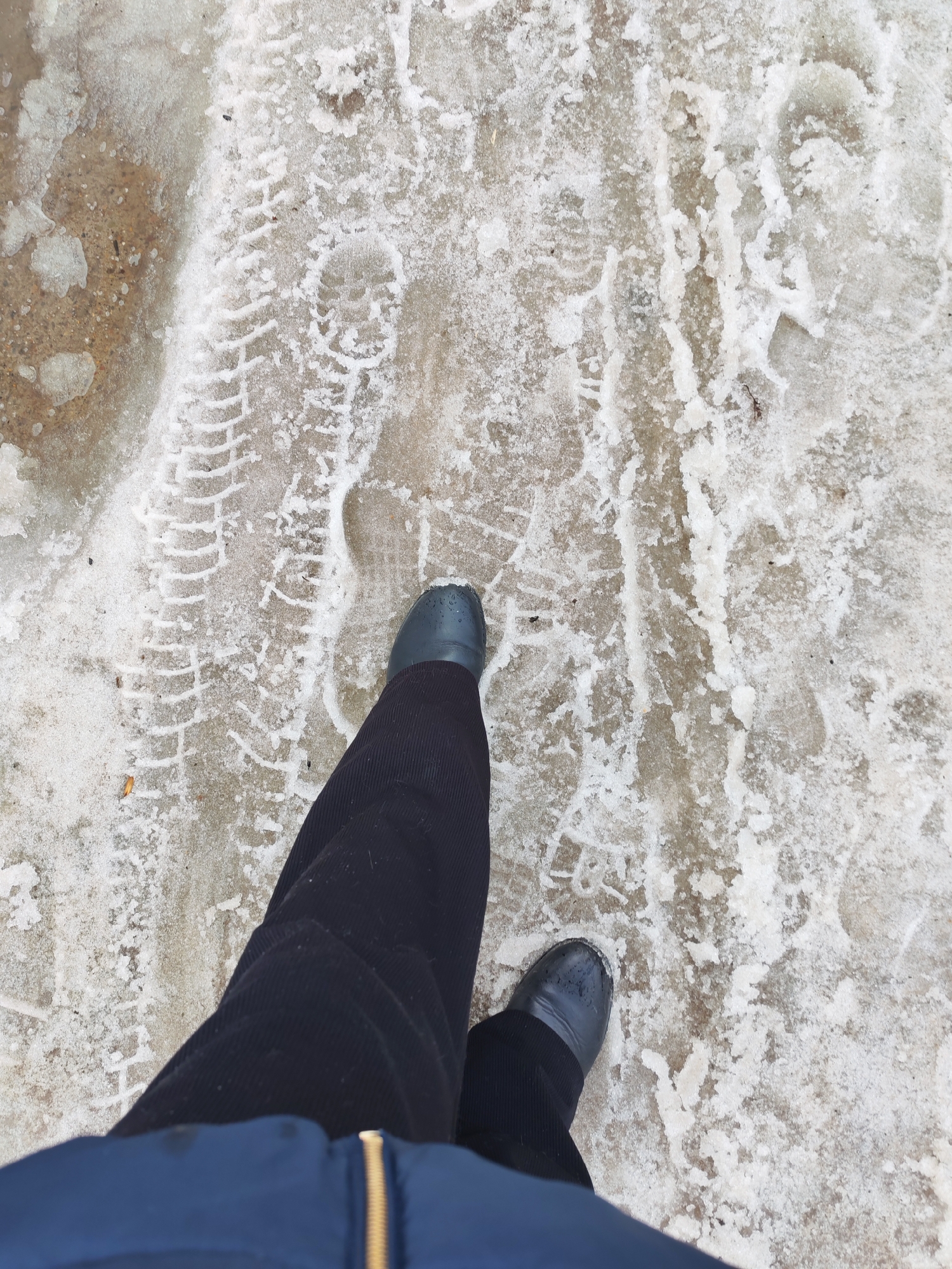 This image, captured in Chaikovsky, Russia, presents a high-angle view of a person's lower body moving across a partially snow-covered, icy outdoor surface. The individual wears dark blue or black ribbed trousers and dark grey boots. The bottom edge of a blue jacket with a visible gold zipper is present in the foreground. The ground surface, likely concrete or asphalt, is mottled with patches of dirty, melting snow and ice, alongside exposed areas of damp pavement. Multiple boot prints and a distinctive tire track pattern are clearly visible within the snow and ice. The subject's right foot is planted on the ground, while the left foot is slightly elevated, indicating a walking motion.