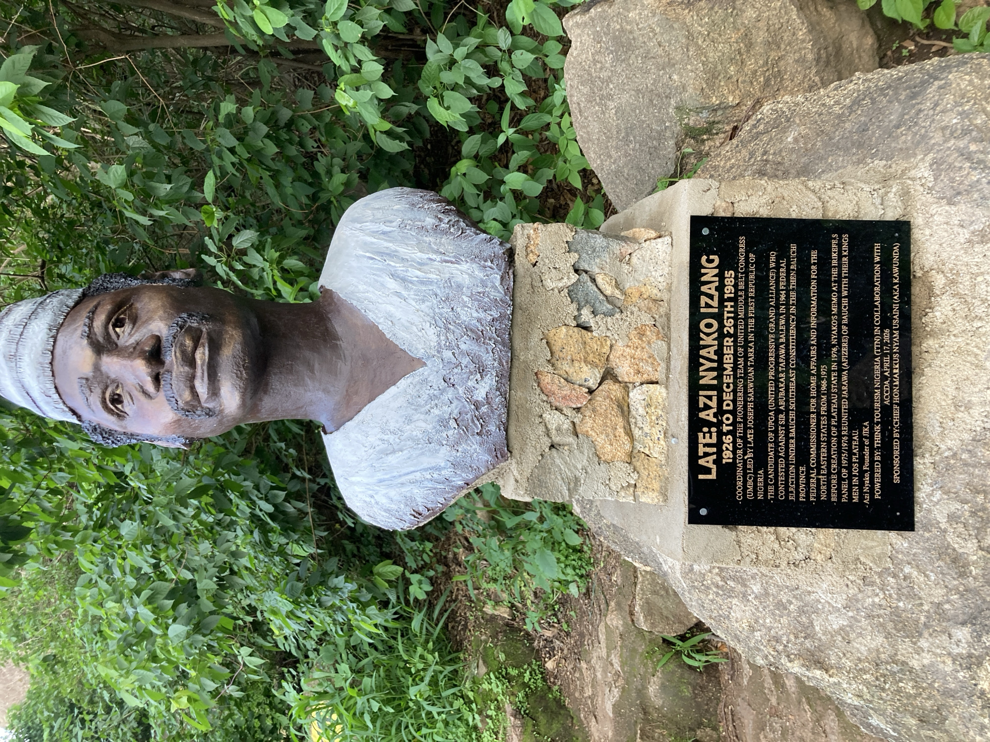 The image shows a bronze bust of a man with a hat, set on a stone pedestal. A black plaque is affixed to the front of the pedestal. The plaque is inscribed with text that includes "LATE: AZI NYAKO IZANG 1926 TO DECEMBER 26TH 1985". The text also lists his achievements and affiliations, such as being the coordinator of the pioneering team of United Middle Belt Congress (UMBC) led by Late Joseph Sarwuan Tarka in the first Republic of Nigeria. It also mentions his role as Federal Commissioner for Home Affairs and Information for the North Eastern States from 1966-1975. The bust and pedestal are situated outdoors, with lush green foliage visible in the background, suggesting a natural or park-like setting. The scene appears to be a memorial dedicated to Azi Nyako Izang.