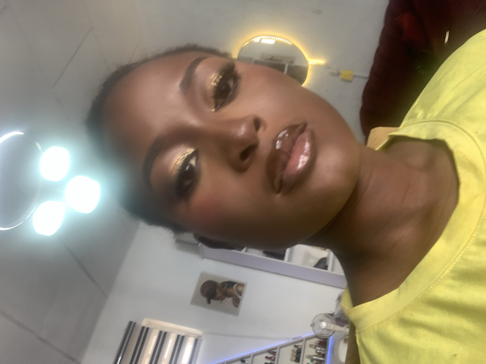 A close-up, high-angle selfie shows a Black woman with dark skin, her face taking up most of the frame. She is wearing bright yellow clothing and has full makeup, including shimmery gold eyeshadow and glossy, plump lips. The background is out of focus, showing a white wall with shelves containing various items, a small fan, and a painting of a woman wearing a baseball cap. A circular mirror with a yellow light ring is visible behind her right shoulder. The lighting in the image is bright, with visible light fixtures overhead casting reflections on the subject's face. The overall impression is of a person taking a selfie in an indoor setting, possibly a dressing room or bedroom. The scene is located in Sabon Tasha, Nigeria.