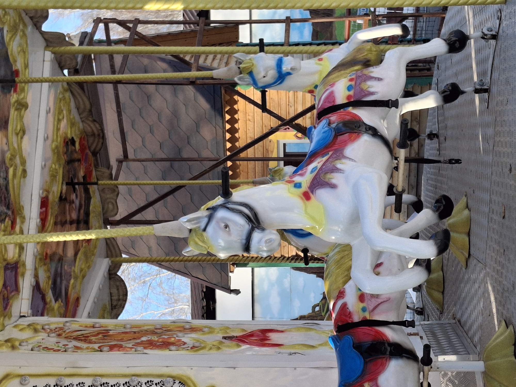 The image shows a carousel with ornate, painted horses. The horses are white with colorful saddles and other decorations. A wooden structure is visible in the background. The carousel appears to be located outdoors, possibly in an amusement park or fairground. The location is Chaikovsky, Russia.