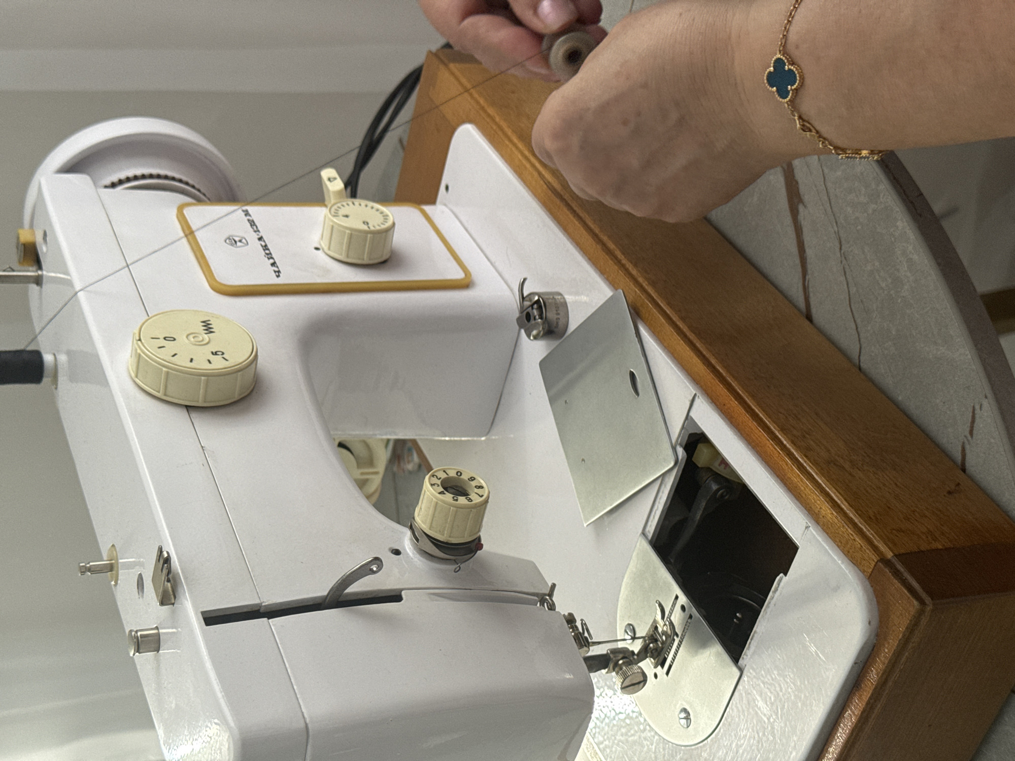 A sewing machine is in focus with a person's hands holding a spool of thread above it. The machine is white with beige dials and a wooden casing. The hands belong to a person wearing a gold bracelet with a dark blue four-leaf clover charm. The sewing machine has the word "ZAGREB" printed on it. The specific location in the city of Saratov, Russia, is not identifiable from the provided image.