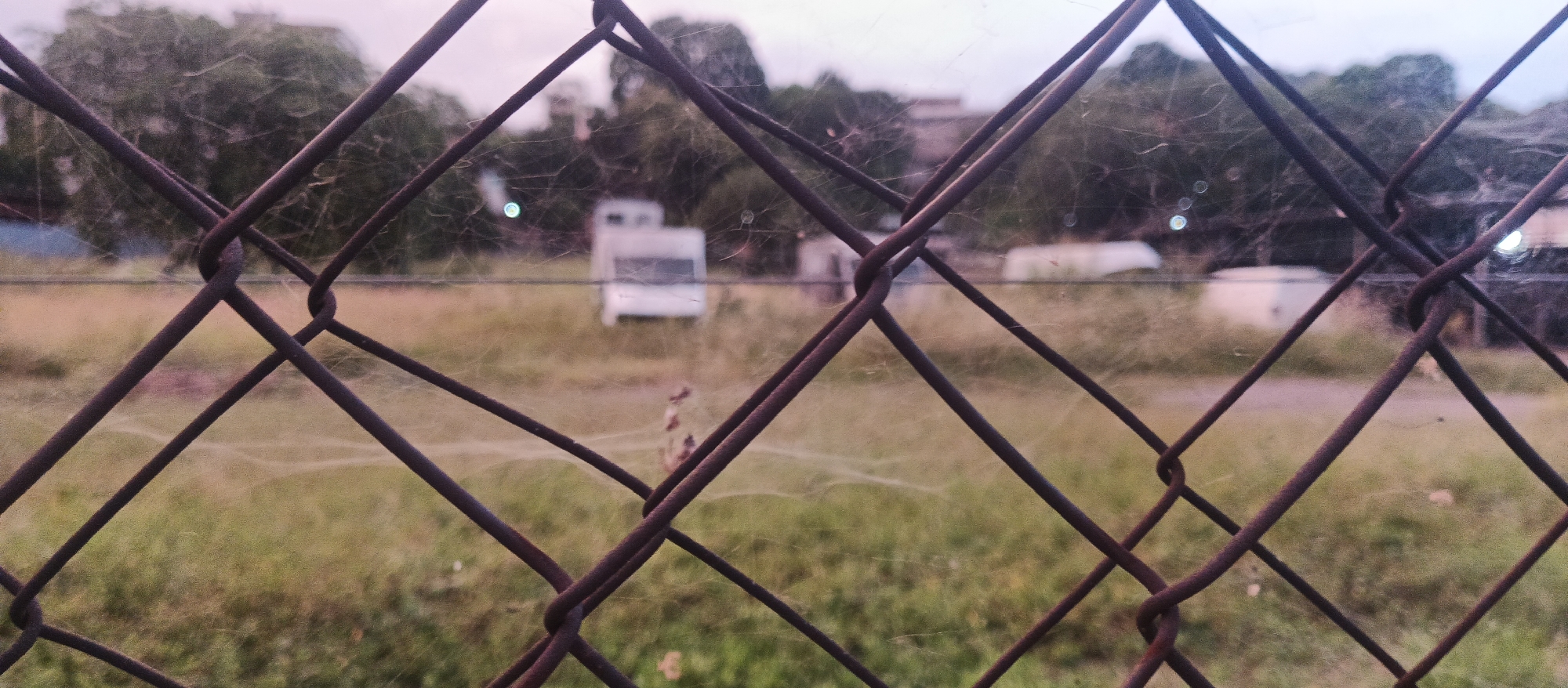 A blurred, out-of-focus image shows a chain-link fence in the foreground. Behind the fence, a grassy field is visible, with some low bushes or weeds scattered throughout. In the middle ground, there are several indistinct white structures, possibly mobile homes or RVs, parked in the field. Trees are visible in the background, with a slightly overcast sky above. The overall impression is one of a somewhat neglected or undeveloped outdoor area.