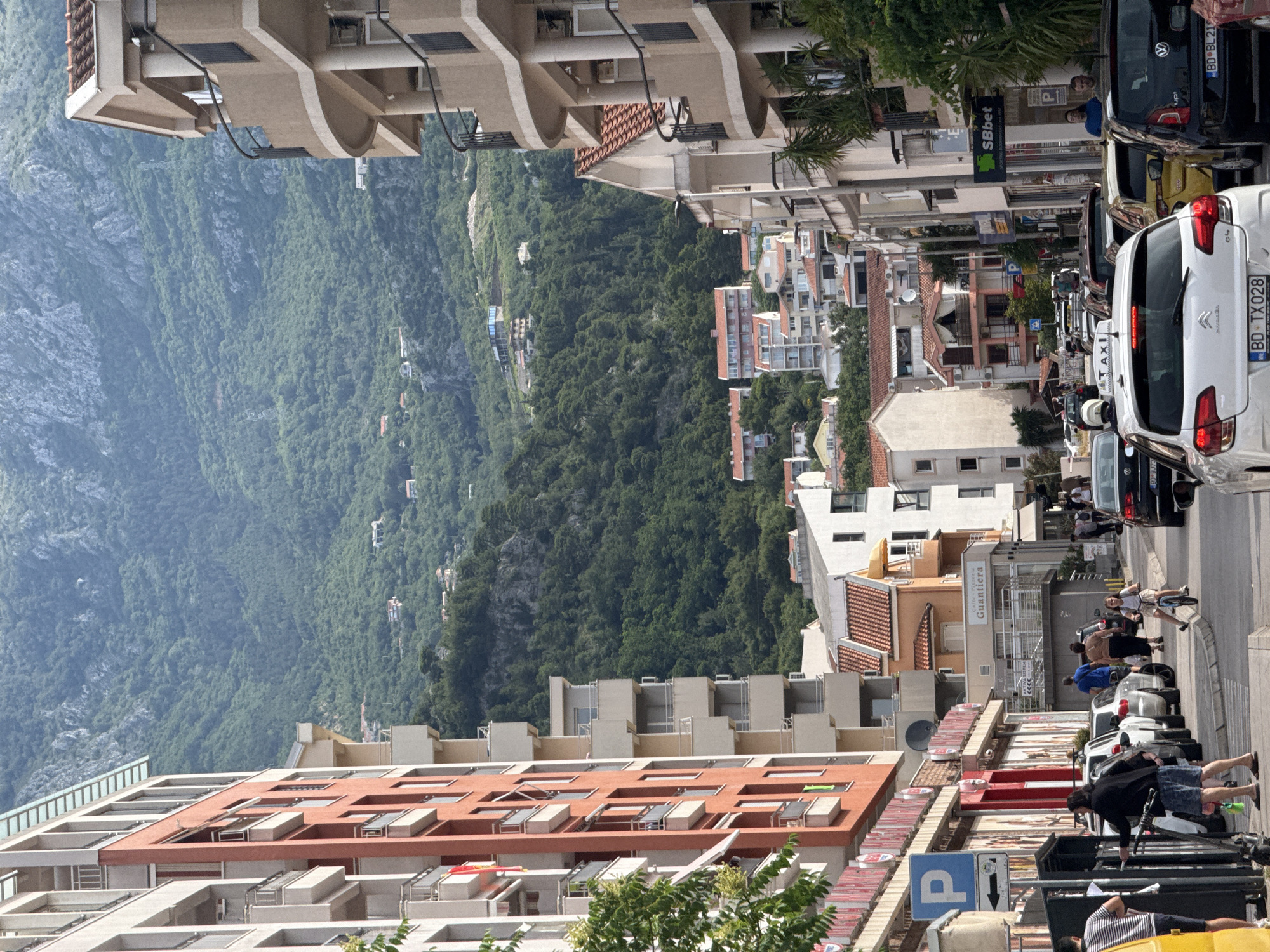 This image, presented rotated 90 degrees clockwise, captures a lively street scene in Budva, Montenegro, set against a dramatic mountainous backdrop. If viewed with the correct orientation, the street extends into the distance, lined with various buildings and a prominent forested mountain dominating the background.
In the foreground, a busy street is visible with numerous parked and moving vehicles. A white Citroën, bearing the license plate "BD TX 028," is prominent on the right, followed by a dark Volkswagen with the plate "BD 80 B219." A "TAXI" sign is also visible on one of the vehicles. Several people are present on the sidewalk and near the cars; one individual is on a scooter or bicycle, while others are standing or walking. A blue "P" parking sign is clearly visible. The buildings along this side of the street are a mix of older, tiled-roof structures and more modern designs. Commercial signs include "SBbet Sportiva Kladionica" and "Caffe Pizzeria Guantiera."
On the opposite side of the street, and extending up the rising terrain, are numerous multi-story buildings. Many display contemporary architectural styles with terraced balconies and flat roofs, likely serving as apartments or hotels. Beyond these urban structures, a vast, densely forested mountain range fills the background, characterized by a lush green canopy. A few smaller buildings are scattered on the lower slopes of the mountain, appearing nestled within the trees.
The scene takes place during the daytime under clear skies, indicating good weather. The overall impression is that of a bustling coastal town, harmoniously blending urban development with stunning natural landscapes.